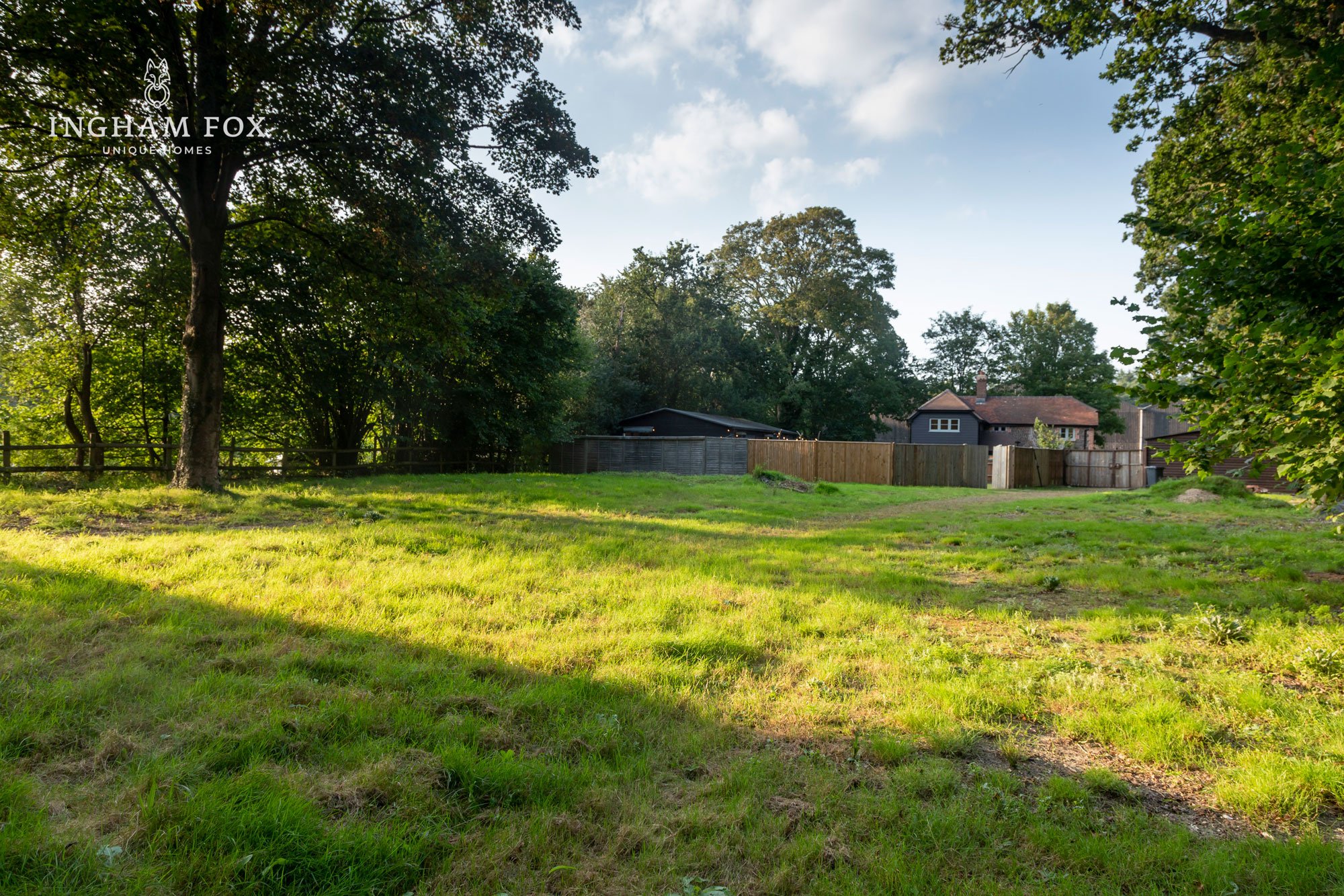 Grassy field with trees, wooden fence, and houses in the background.