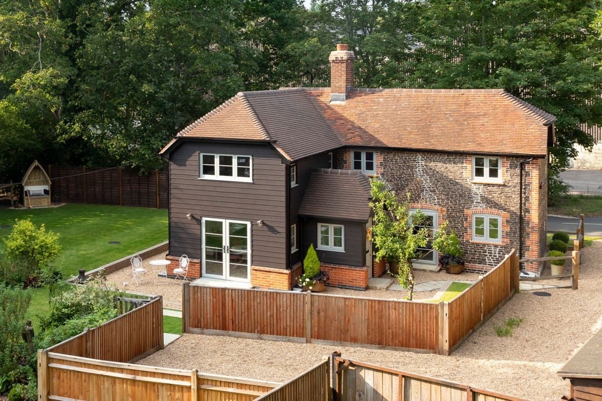 A charming two-story house with a brick and wood exterior, surrounded by a wooden fence and a lush green garden. The house features a pitched roof, large windows, and a small patio area.