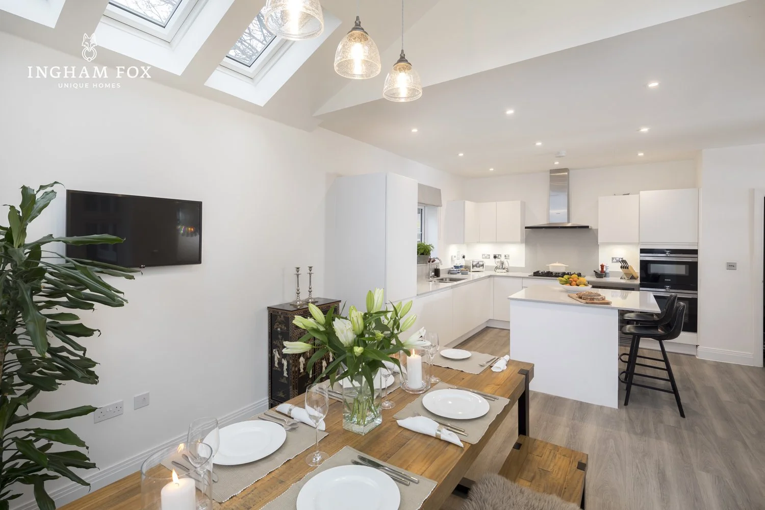 Modern kitchen and dining room with skylights, white cabinets, a set dining table with plates and glasses, a plant, and a black wall-mounted TV.