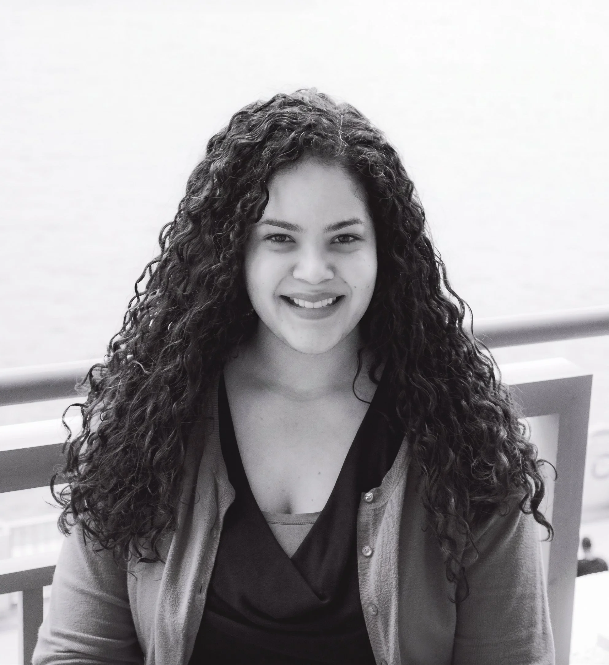 Close-up black and white photo of a woman with curly hair smiling, wearing a cardigan over a shirt, sitting in front of a white railing with horizontal slats.