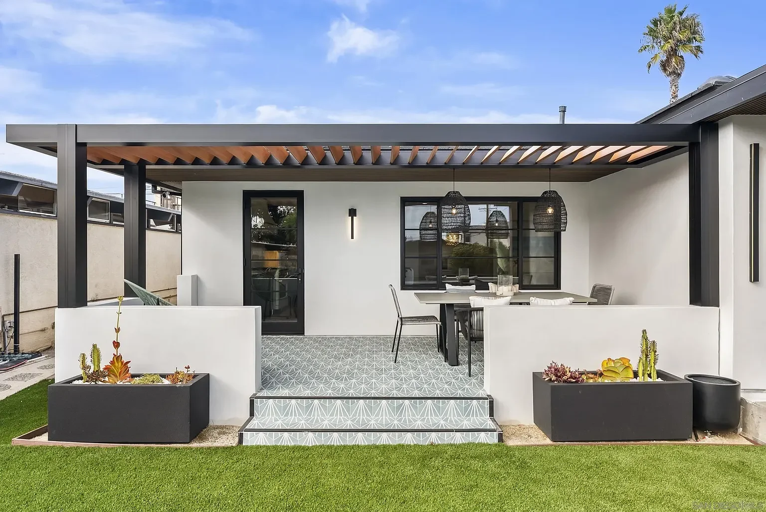 Modern outdoor patio with black and white furniture, hanging lanterns, potted succulents, and a covered roof, overlooking a green lawn.