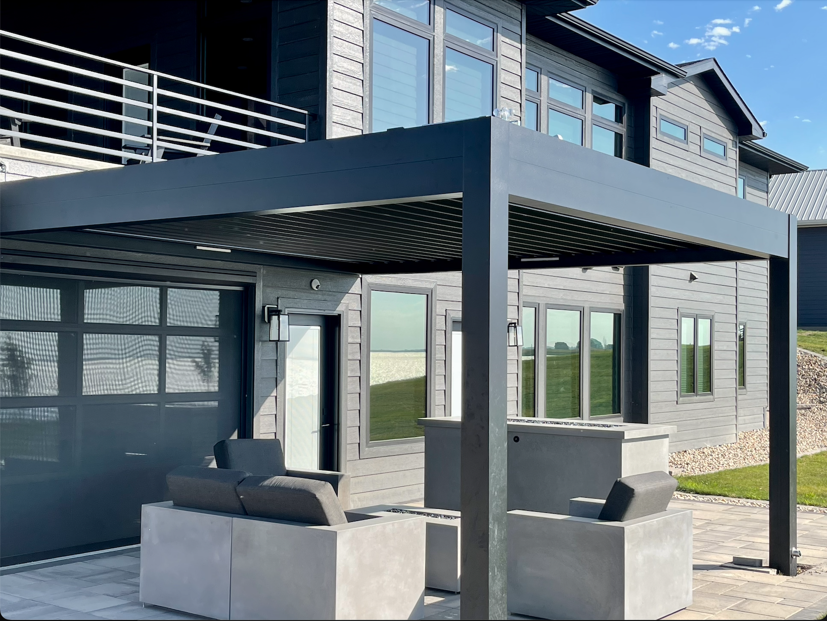 Modern house exterior with a metal pergola and outdoor seating area. The house features large windows and a second-story balcony with railings. The patio has cushioned gray seating and a stylish minimalist design. A manicured lawn and clear blue sky 