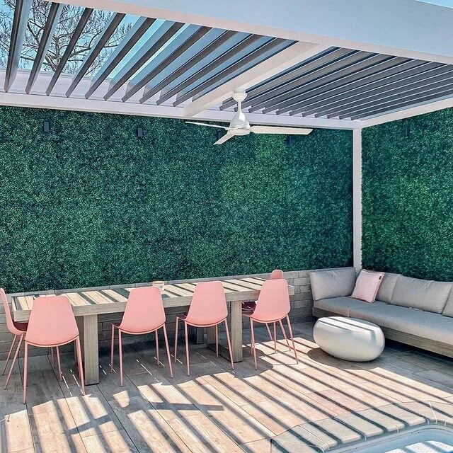 Outdoor patio with pink chairs, modern dining table, green hedge wall, gray sectional sofa, round white ottoman, and a pergola ceiling with fan.