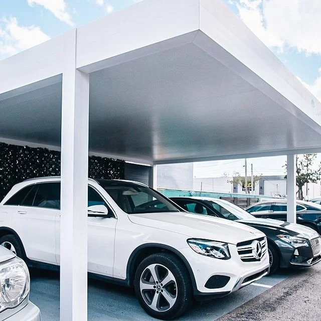 Cars parked under a modern white shelter, including a white SUV, in an outdoor setting on a sunny day.