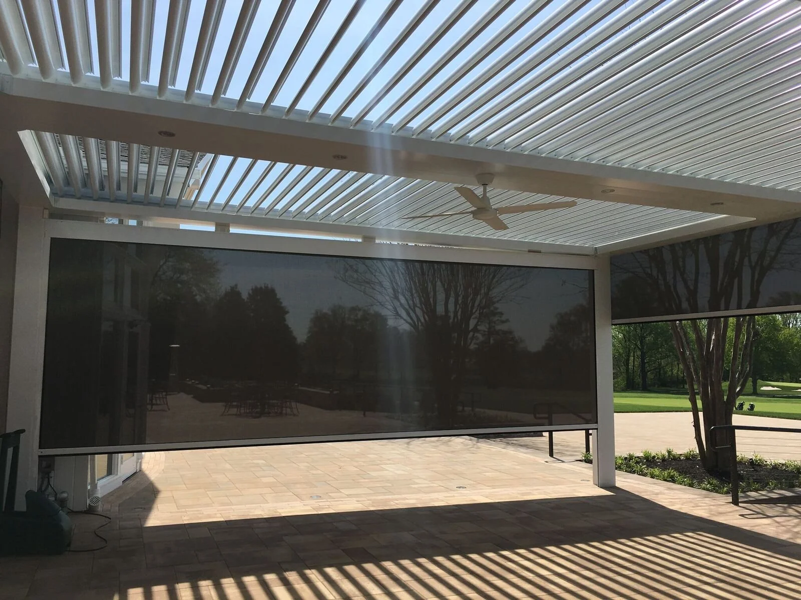 Patio with screened enclosure, ceiling fan, and sunlight casting shadows on the tiled floor, overlooking a park with trees and grass.