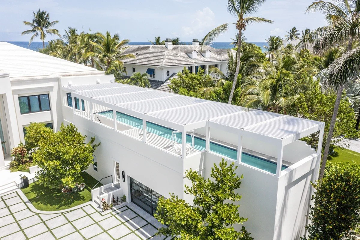 Aerial view of a modern luxury home with a rooftop lap pool, surrounded by palm trees and tropical vegetation. The house has a sleek, white exterior with a large driveway. Ocean can be seen in the background.