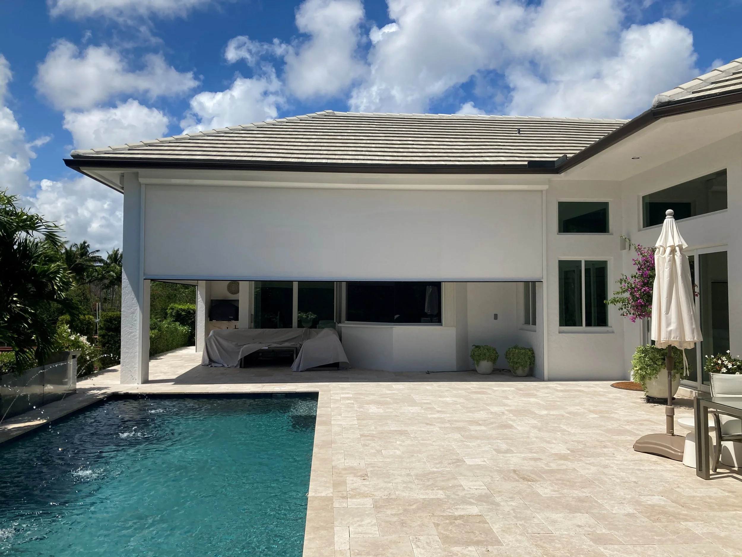 Modern home exterior with a pool, tiled patio, an outdoor shade, and a closed umbrella beside a table and chairs. Bright, clear sky in the background.