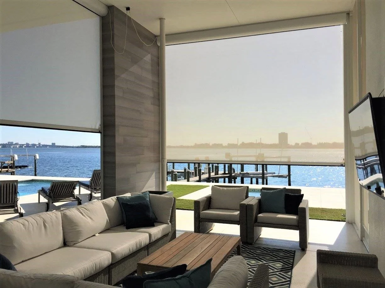Modern outdoor patio with comfortable seating overlooking a waterfront view, featuring couches and chairs, a coffee table, and a flat-screen TV.