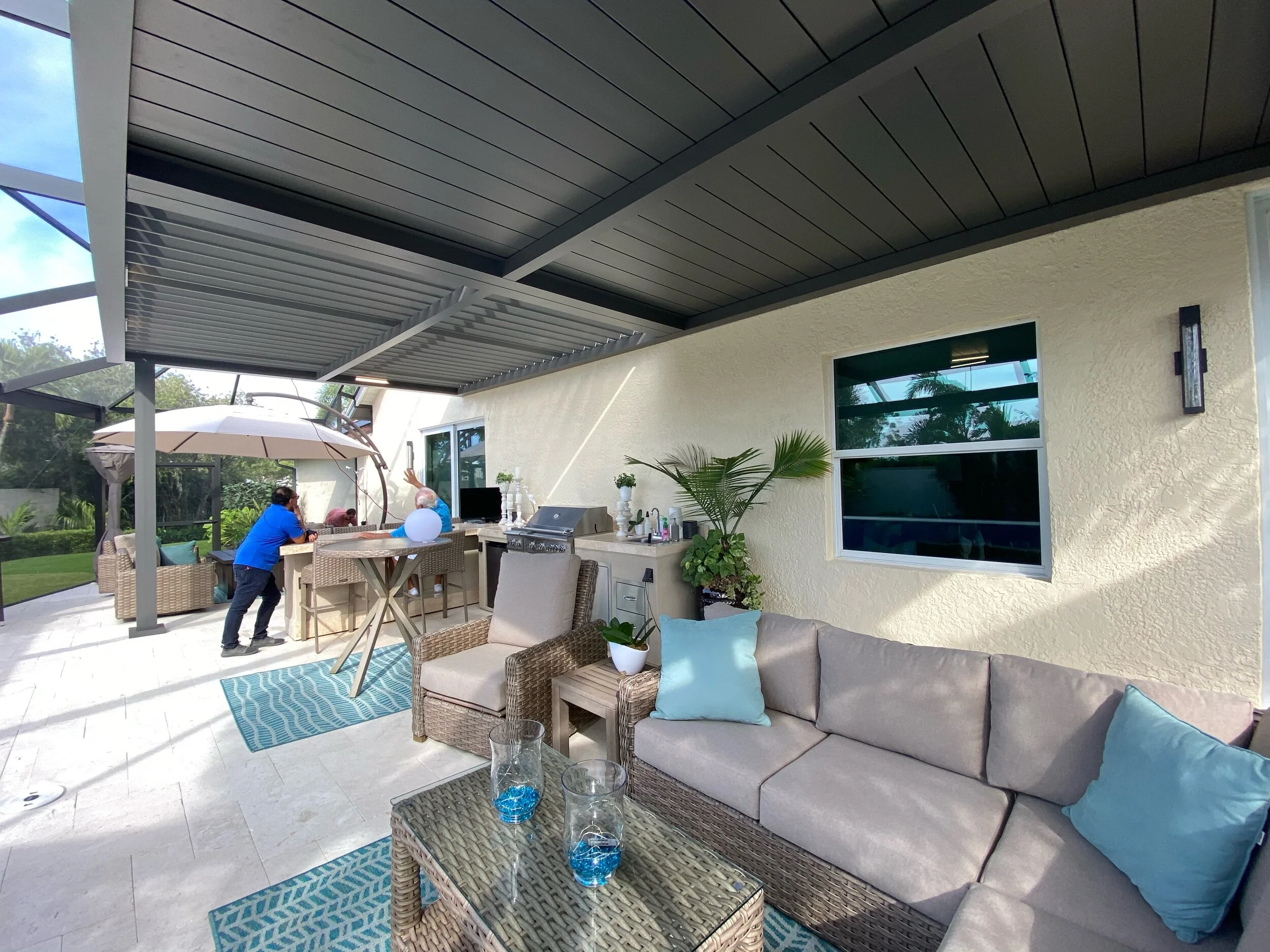 Outdoor patio with comfortable seating, a covered pergola, and decorative elements like blue cushions and mats. Two people are at a bar area, with plants and greenery in the background.
