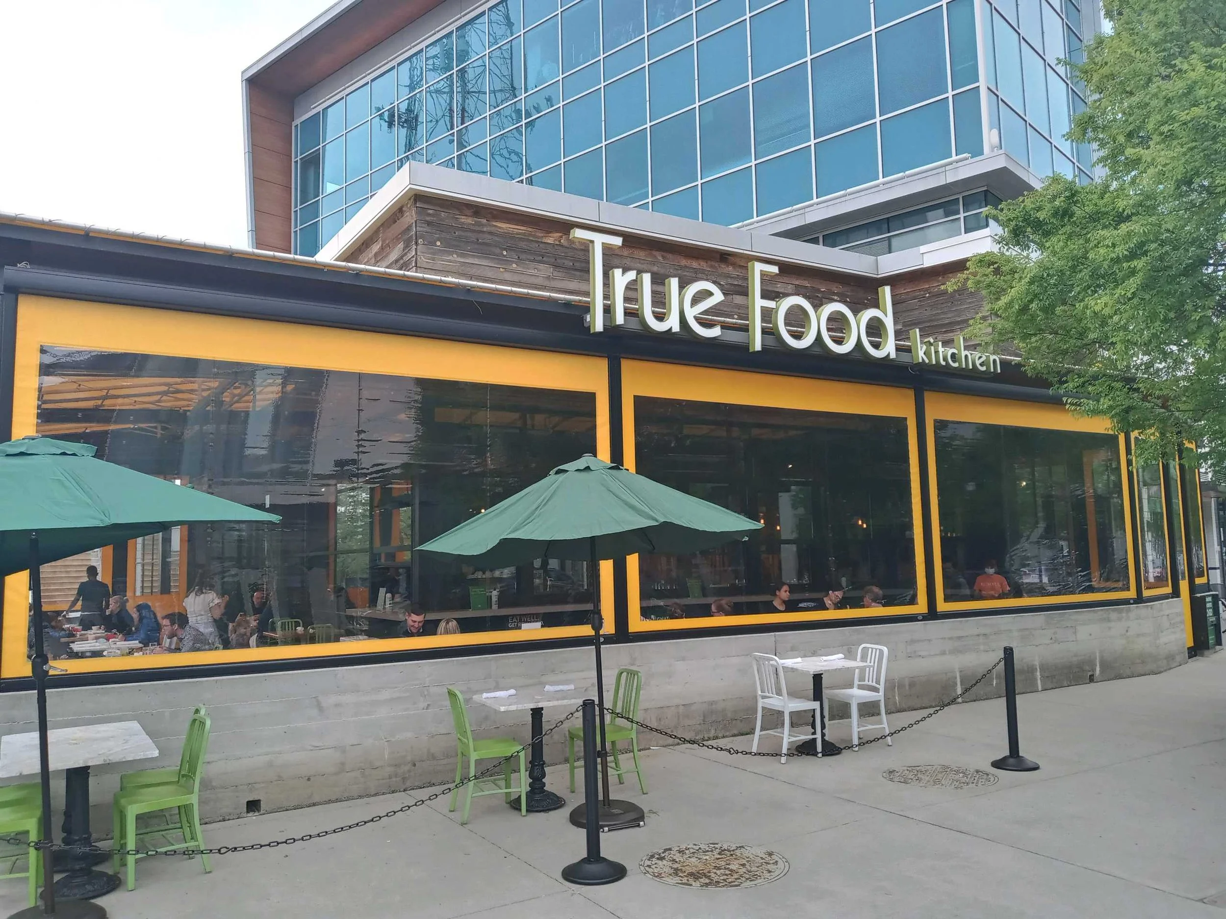 Exterior of True Food Kitchen restaurant with outdoor seating, green umbrellas, and glass windows showing diners inside.