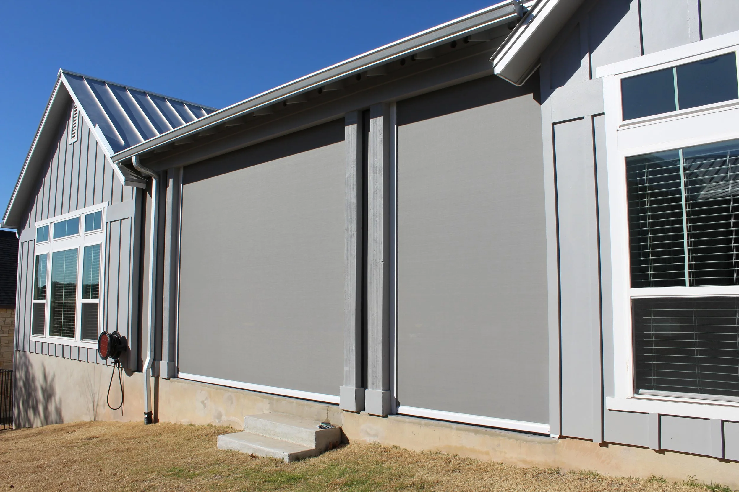 Modern house exterior with large window shades and metal roof, featuring a light gray color scheme and external blinds partially covering windows.