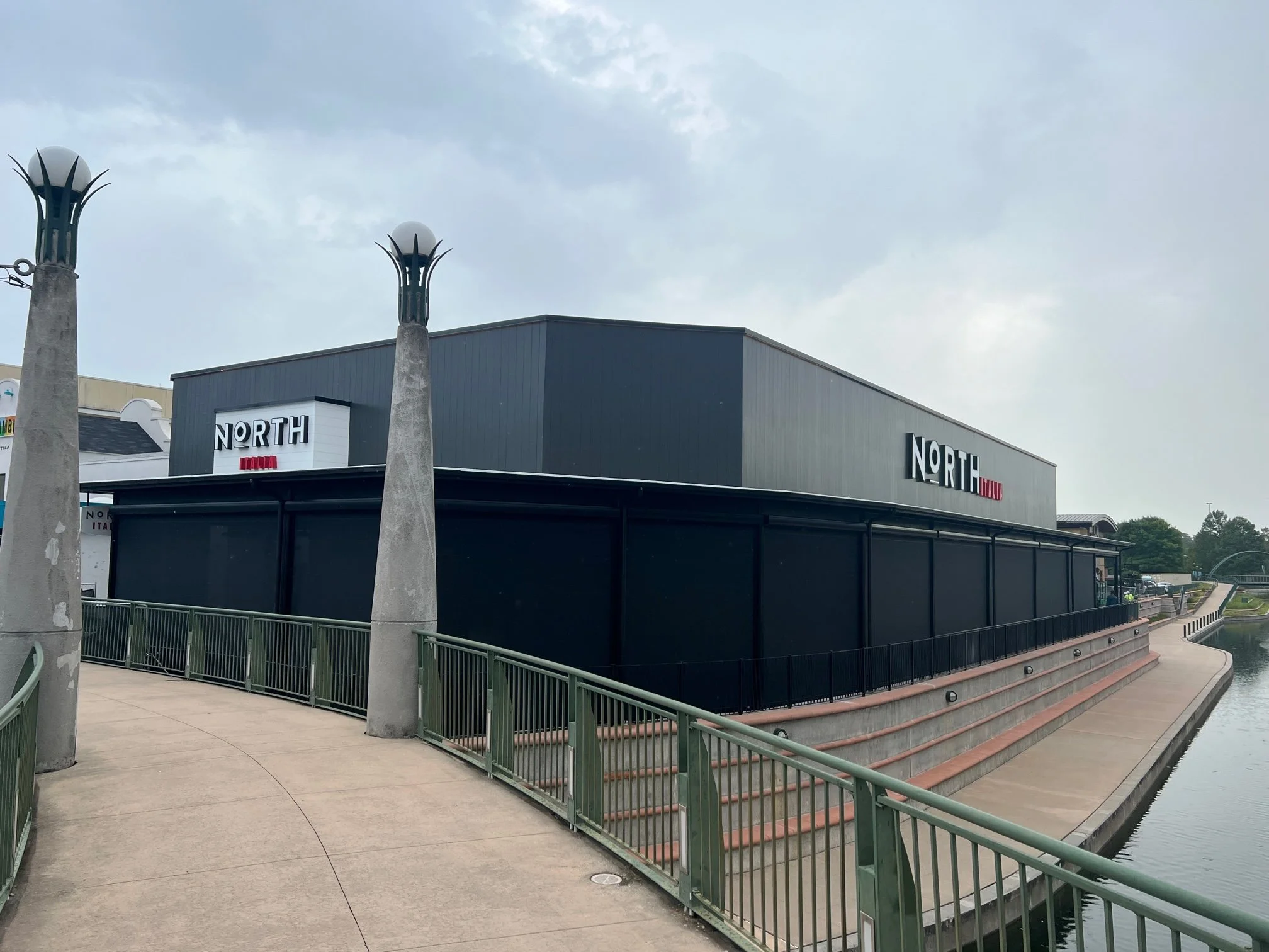 Exterior view of the "North Italia" restaurant with modern architectural design, adjacent to a walkway and railing. The building features large block letters for signage and is positioned by a waterway.