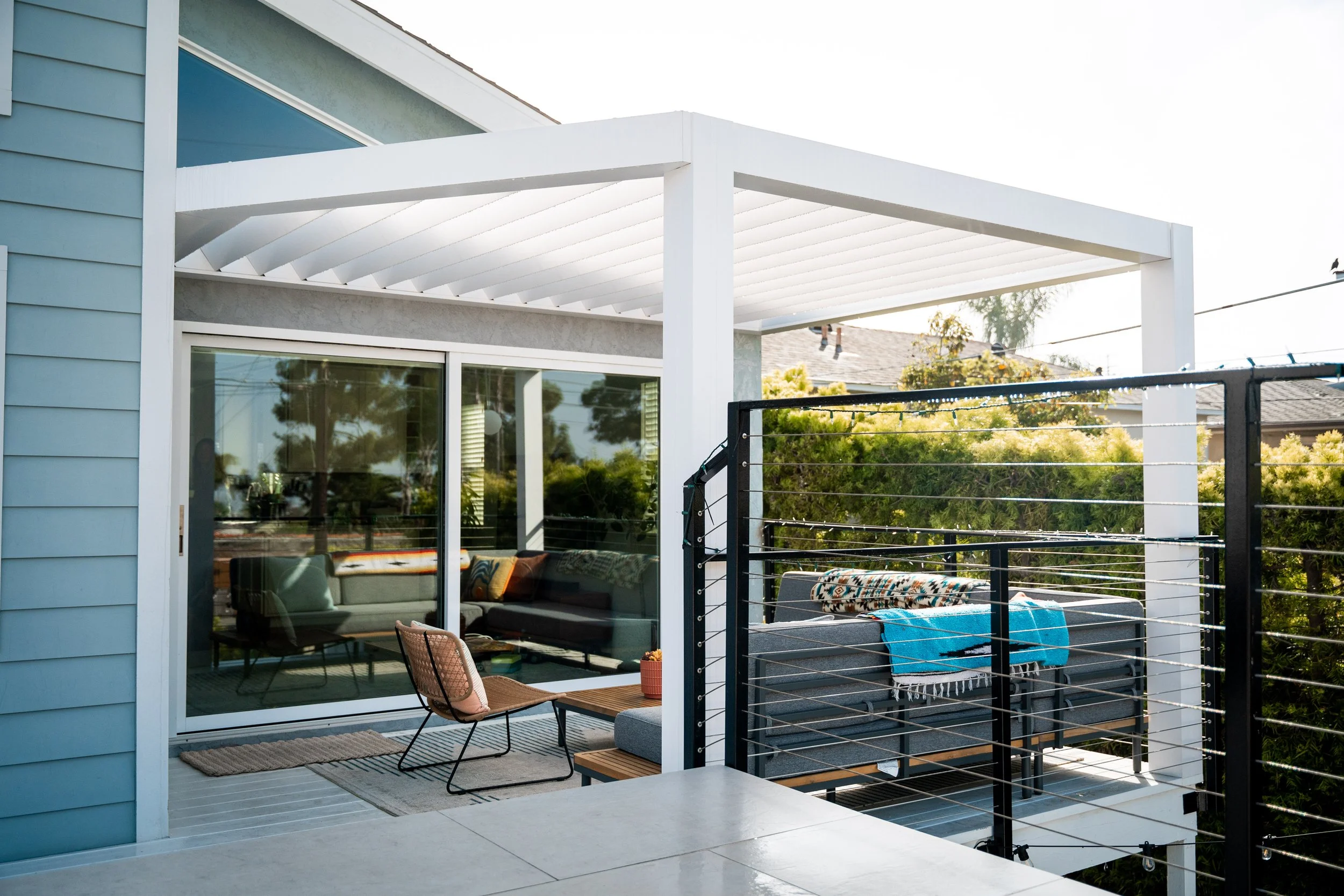 A balcony with outdoor furniture, including a chair, tables, and cushions, overlooking trees and neighboring houses, with a modern house exterior and a white pergola above.