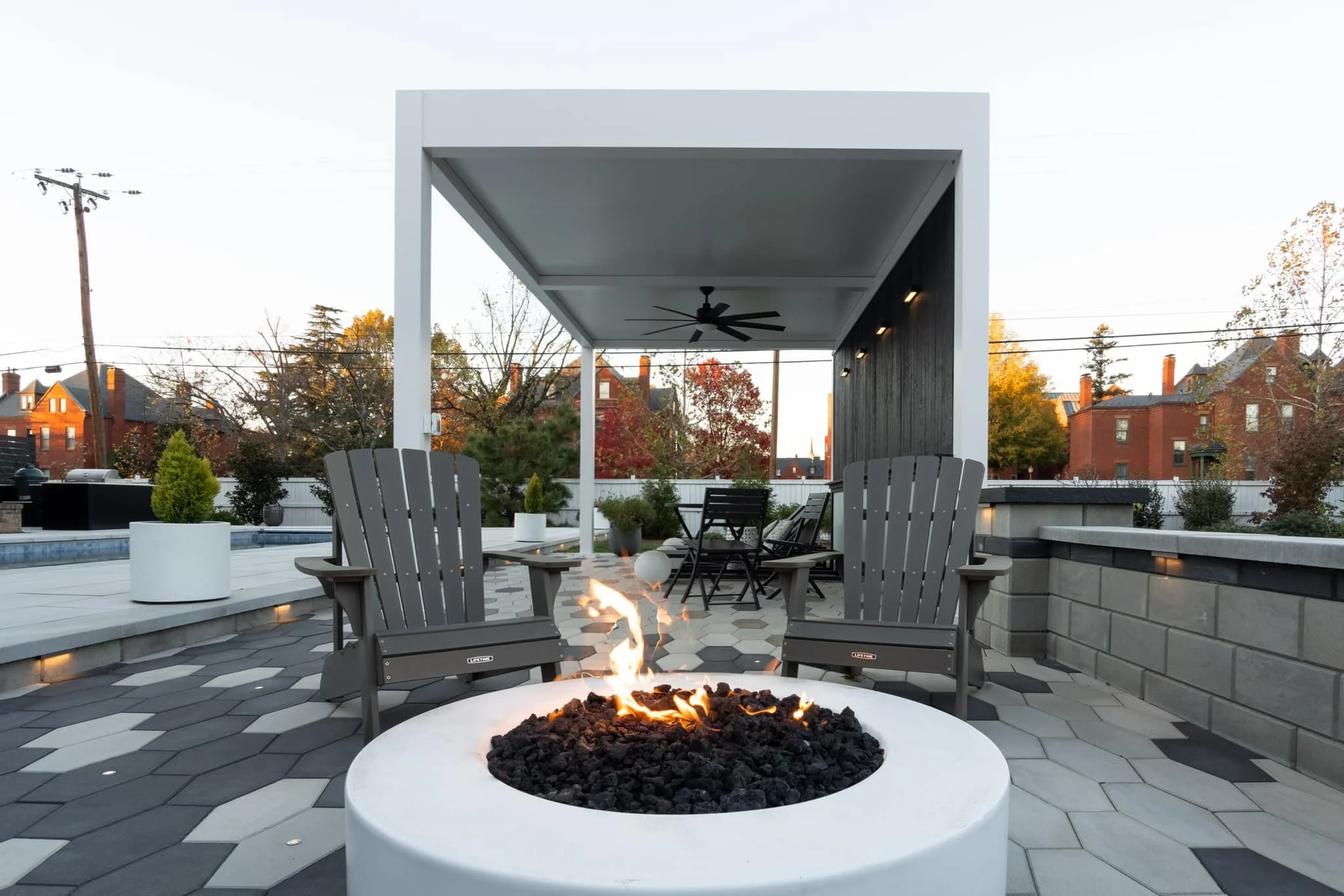 Outdoor patio with a modern fire pit and Adirondack chairs under a pergola, surrounded by geometric paving tiles and landscaped greenery, with residential homes in the background.