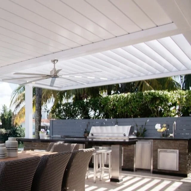Modern outdoor kitchen with white pergola, ceiling fan, dining table, wicker chairs, built-in grill, and lush greenery.