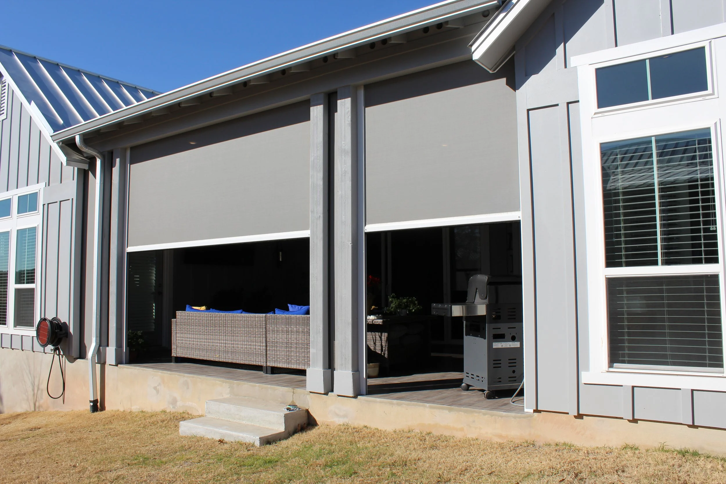 Outdoor patio with retractable shades, wicker furniture, grill, and a modern house exterior.