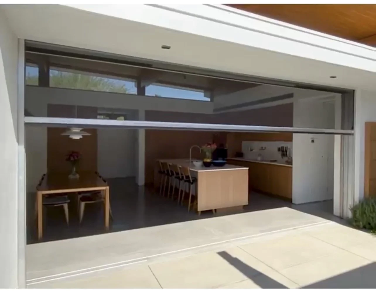 Modern kitchen with open sliding glass door, revealing a dining area with a wooden table and chairs, a kitchen island with bar stools and motorized shade screens.