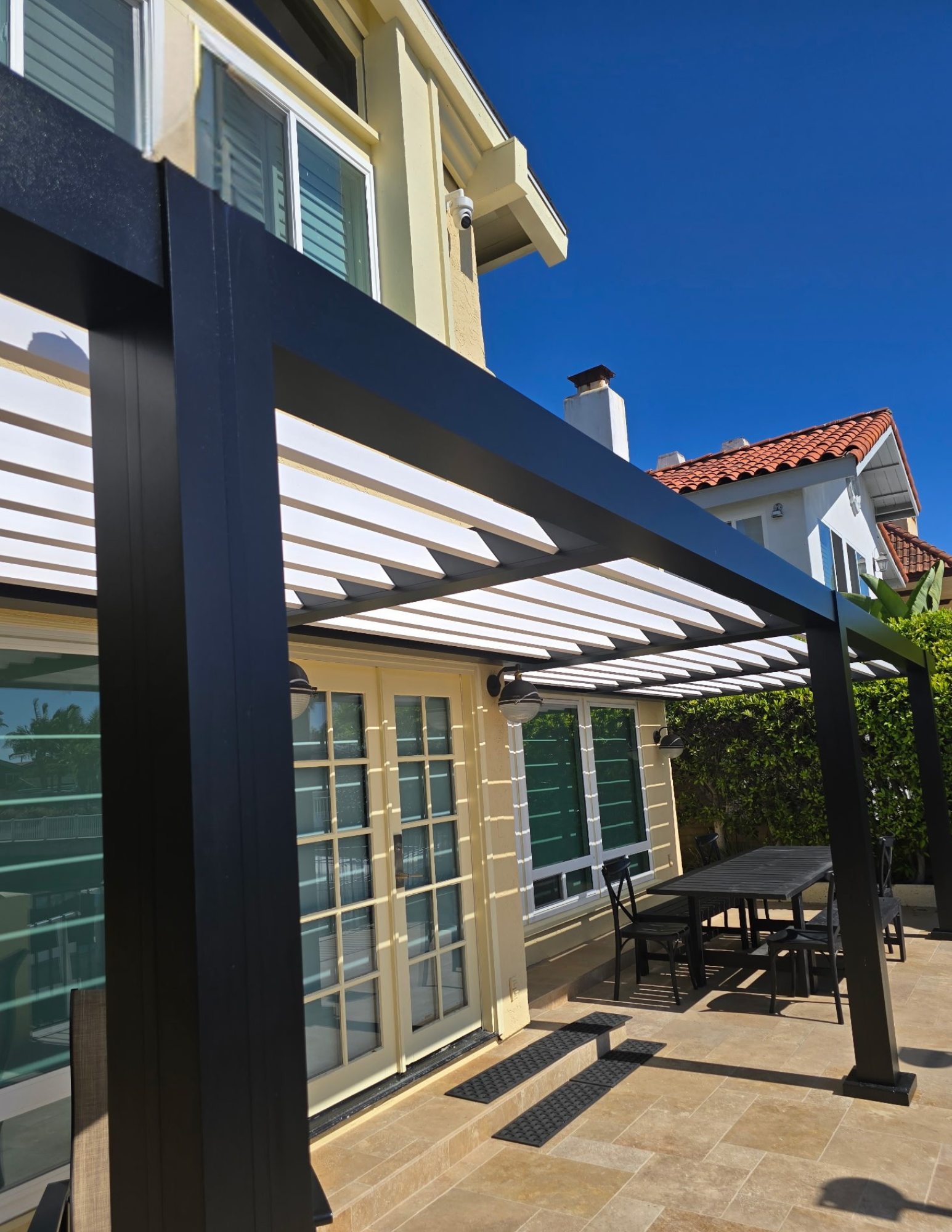Outdoor patio with a black and white pergola, patio furniture including a table and chairs, and a house with large sliding glass doors and windows under a clear blue sky.