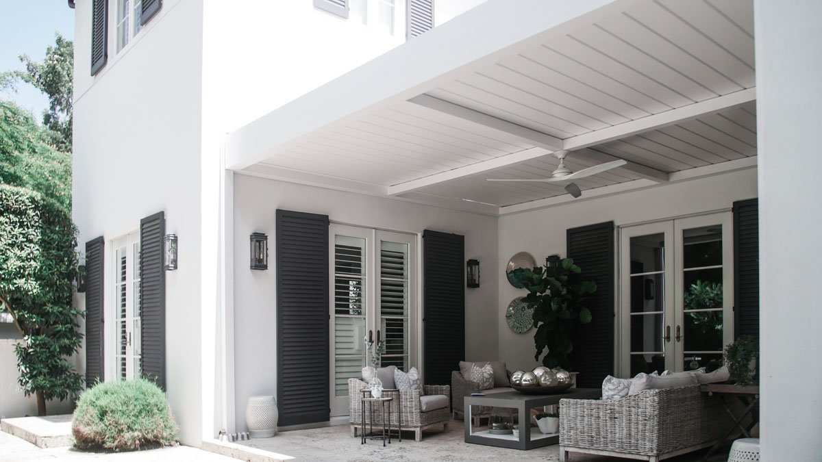 Motorized modern louvered patio with wicker furniture, white walls, black shutters, ceiling fan, and potted plants.