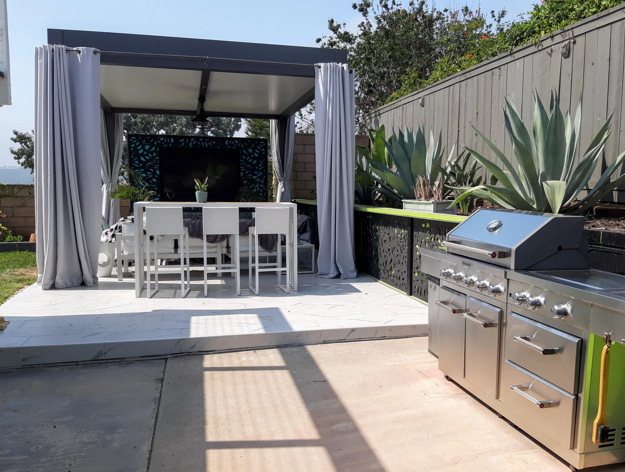 Modern outdoor patio with a pergola, dining set, and stainless steel grill. Surrounding the area are large green agave plants and gray privacy curtains. A flat screen TV is mounted in the background.