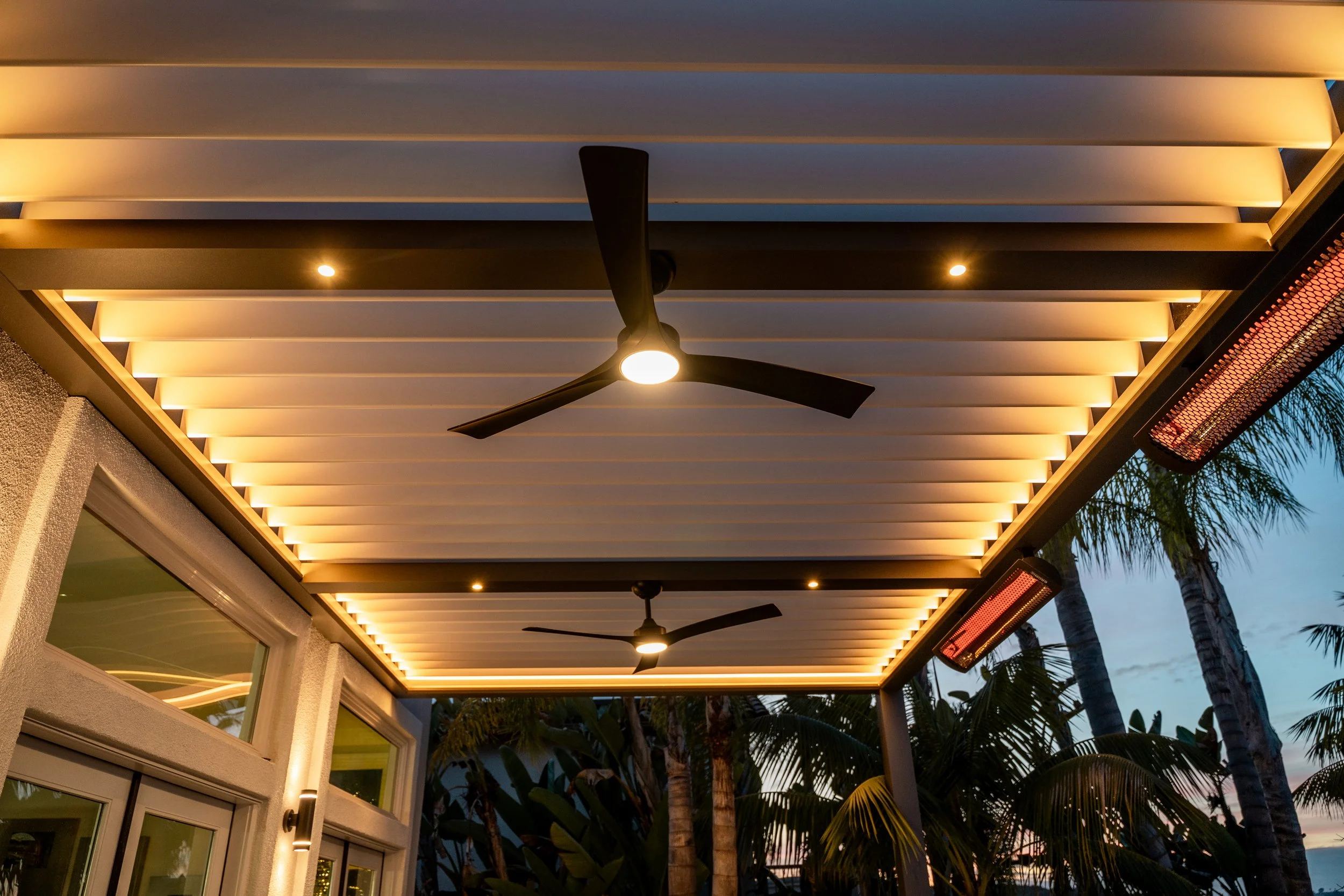 Motorized modern outdoor patio with ceiling fans, LED lighting, and infrared heaters, surrounded by palm trees in San Diego, CA.