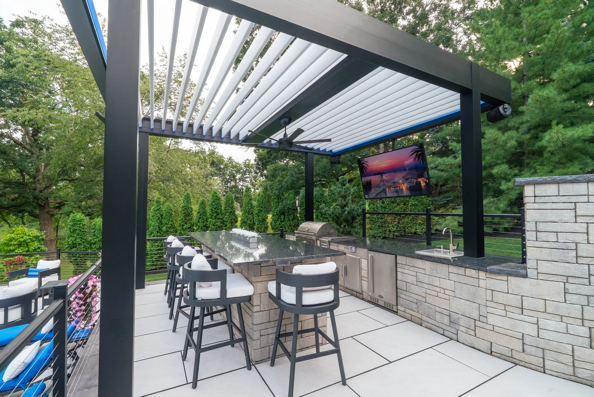 Outdoor patio with a pergola featuring adjustable slats, ceiling fan, mounted television showing a sunset beach scene, and bar seating area surrounded by lush green trees and plants.