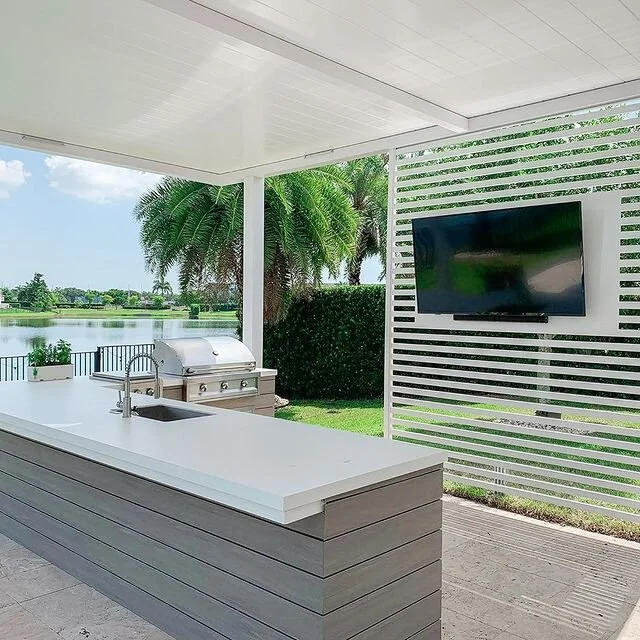 Modern outdoor kitchen with countertop, sink, and grill overlooking a lush garden and lake, featuring a mounted TV on a slatted white wall under a covered patio.