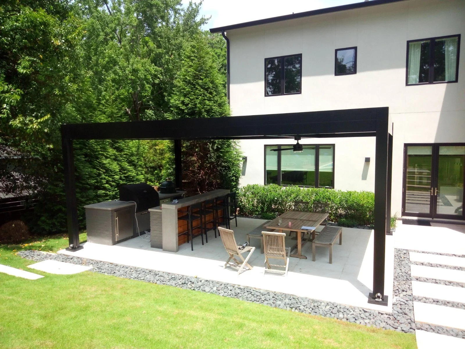 Modern outdoor patio with a canopy, featuring a kitchen area with a grill and bar counter, and a dining space with a wooden table and chairs, set on a stone-paved area, surrounded by green lawn and hedges.