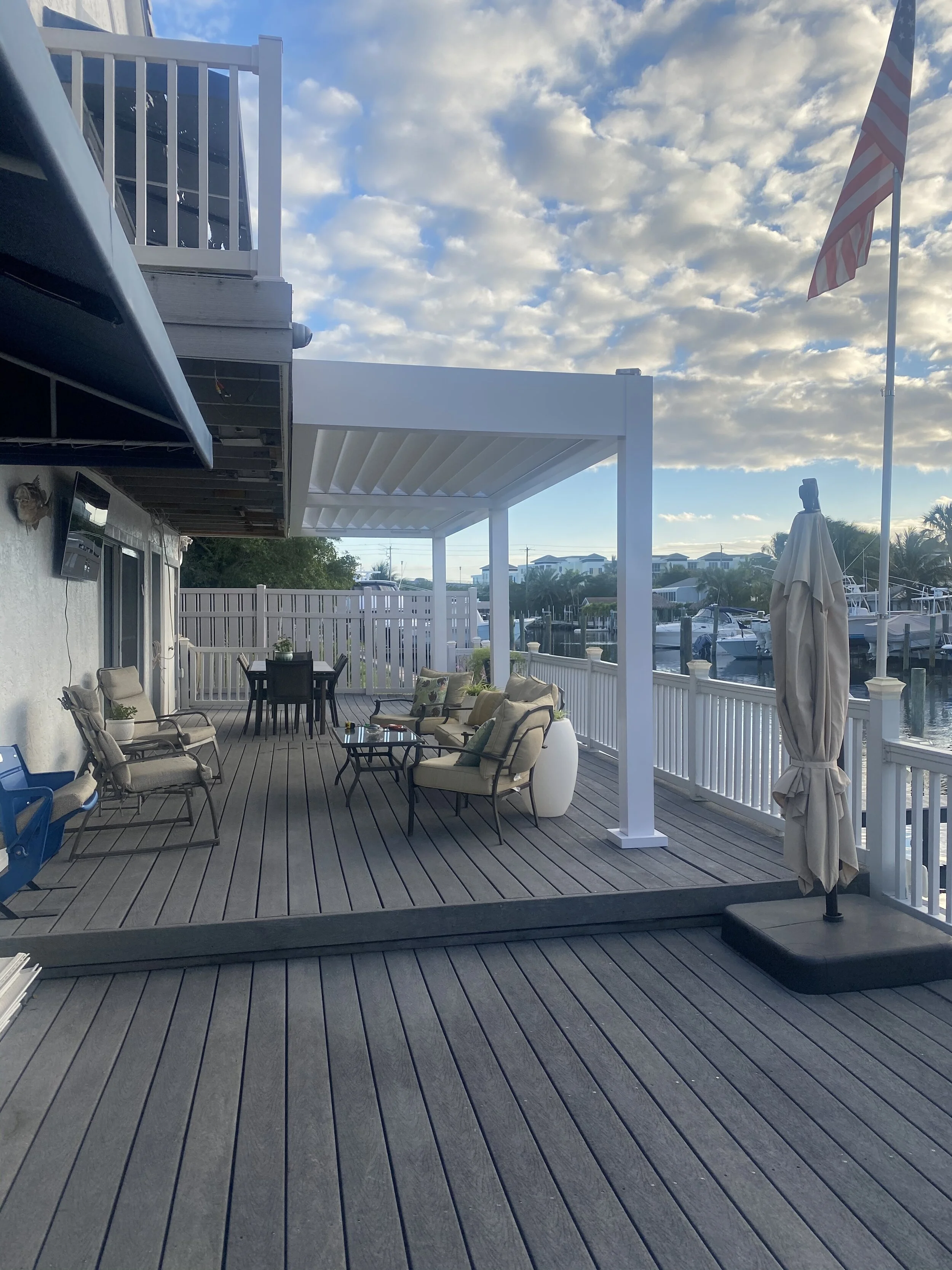 Outdoor patio with cushioned chairs, a dining table, and a pergola, overlooking a canal. An American flag is visible on a pole. The area is surrounded by a white railing and cloud-filled sky.