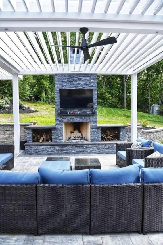 Outdoor patio with wicker furniture and blue cushions under a pergola, featuring a stone fireplace with a mounted TV and a ceiling fan.