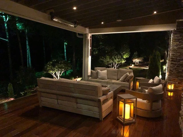Indoor outdoor living area with couches, armchairs, lantern-style lights, and a view of a dark outdoor landscape with trees at night.