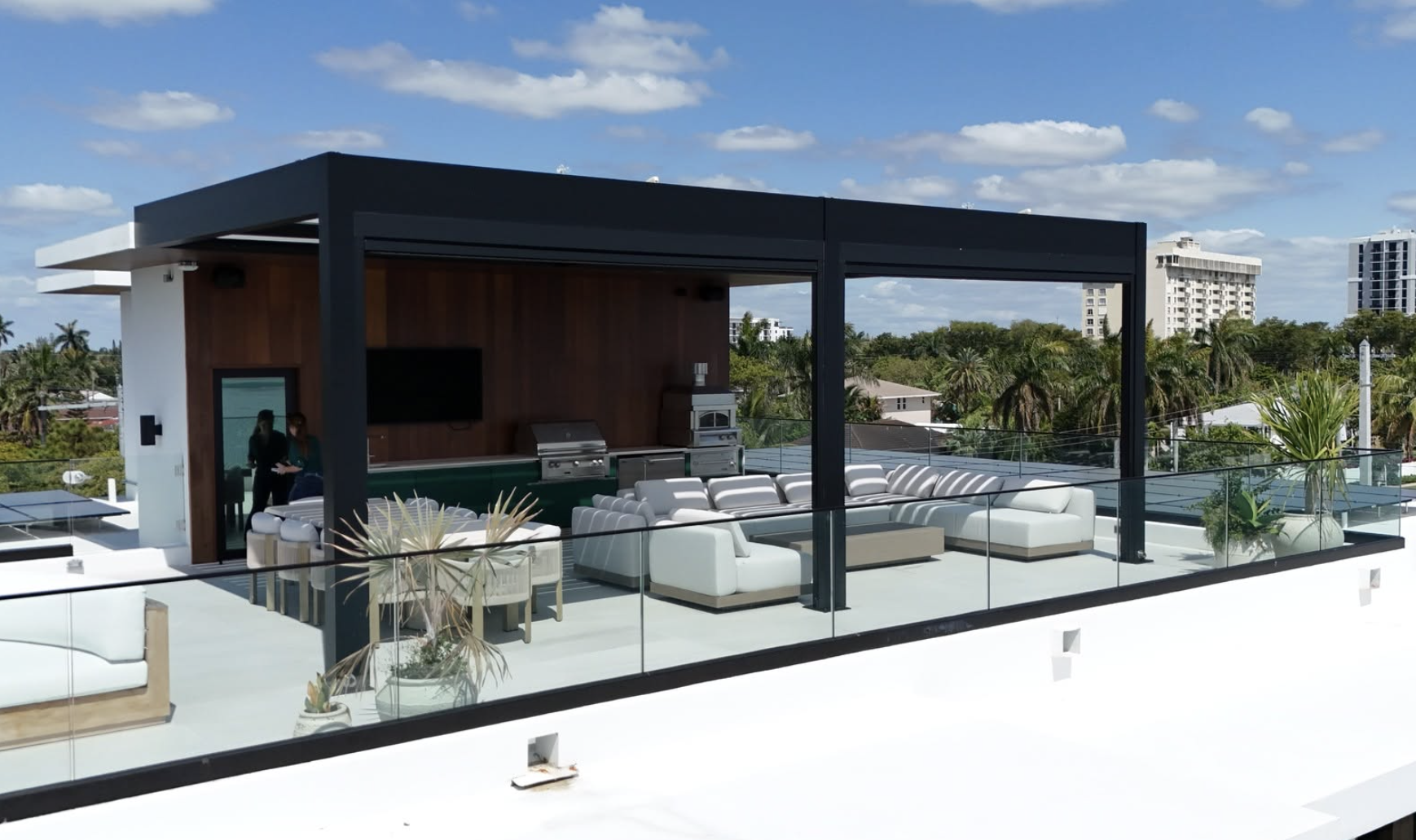 Rooftop outdoor living area with white sectional sofas, potted plants, a built-in grill, and a mounted TV under a black modern pergola on a sunny day with a blue sky and white clouds.