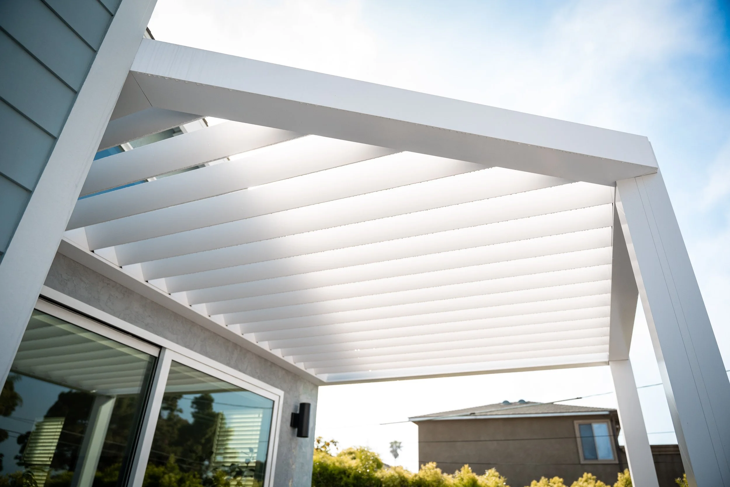 A white pergola attached to a house with a sliding glass door, overlooking other houses and trees in the background.