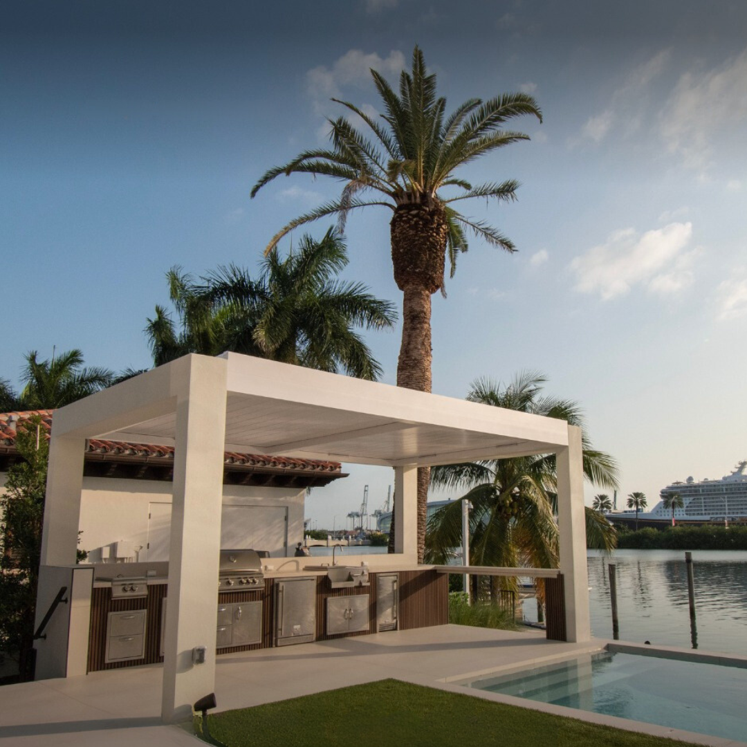 Outdoor kitchen with pergola, palm trees, and pool beside a waterfront.