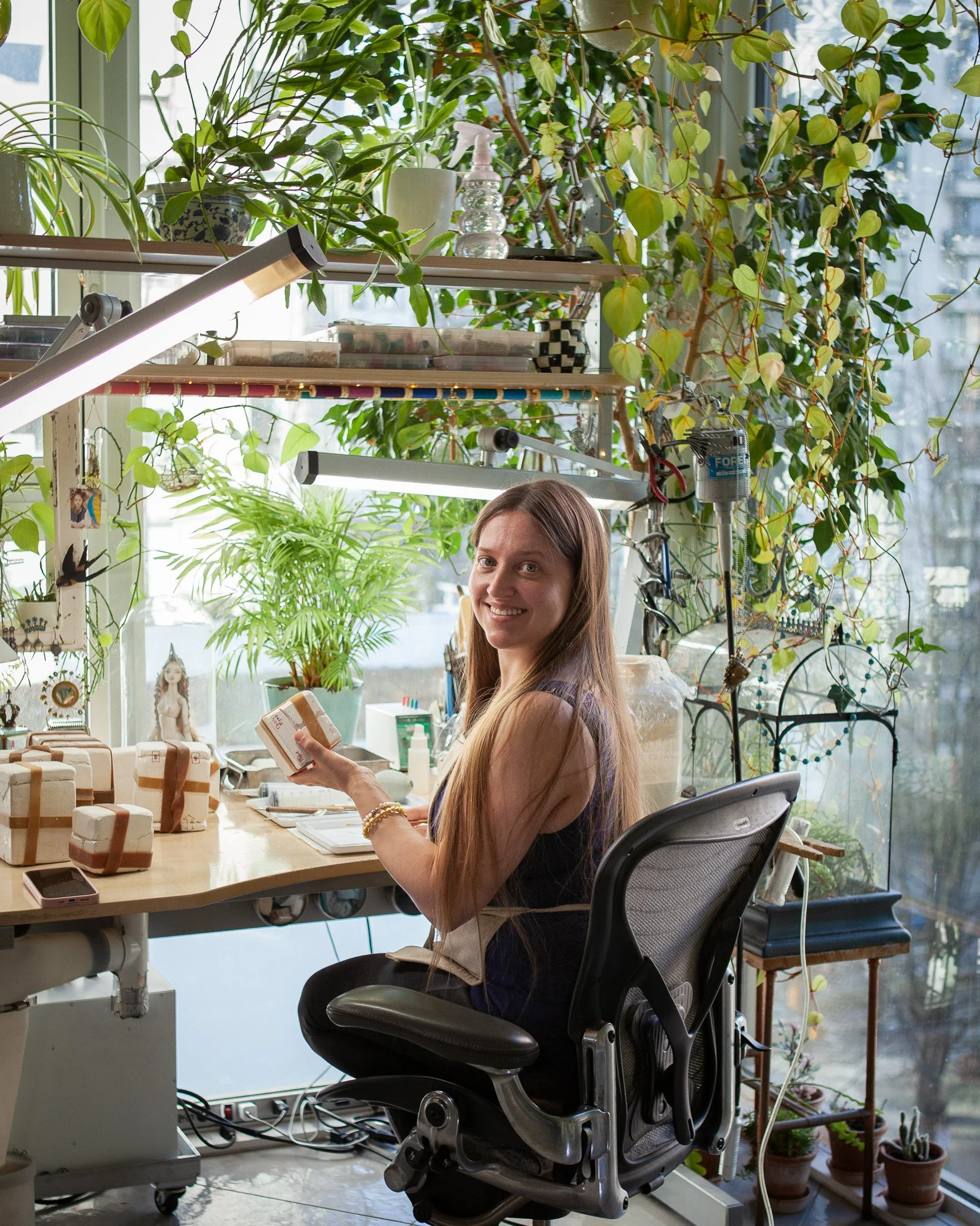 Marina Bychkova sitting at her desk in her studio, working with porcelain doll molds, surrounded by plants