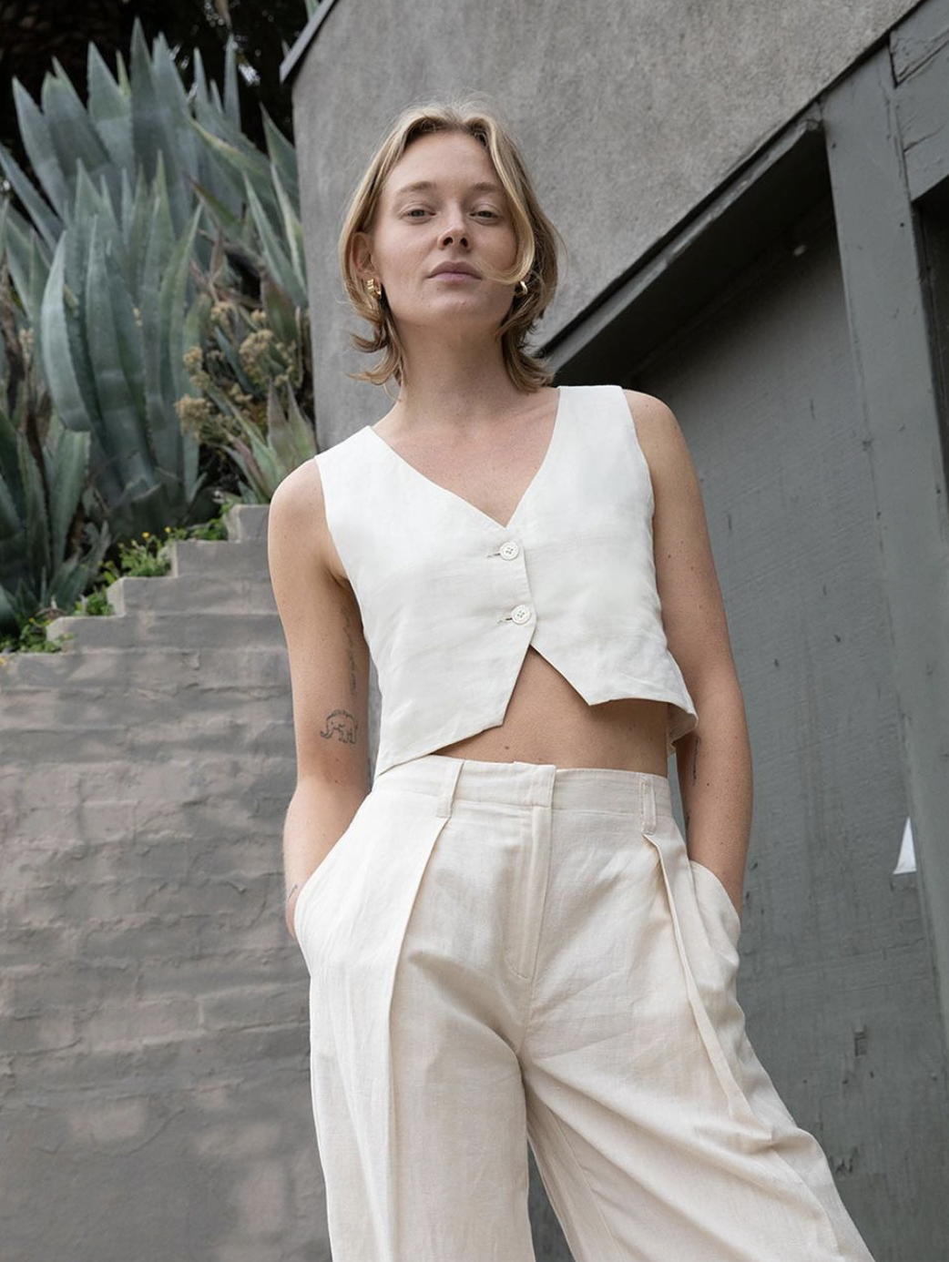 A woman with short light brown hair wearing a sleeveless white crop top with buttons and high-waisted cream-colored pants, standing outdoors in front of plants and a house with gray walls.