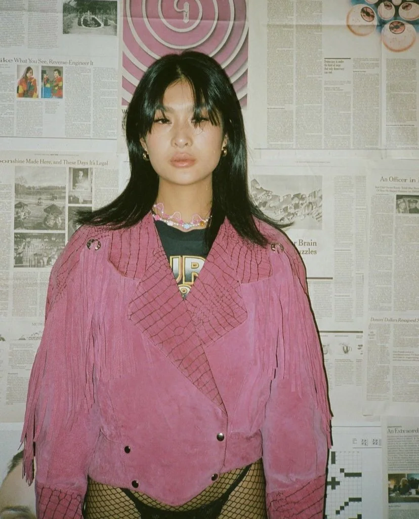 A woman with black hair, wearing a pink fringed jacket and fishnet stockings, standing in front of a wall covered in newspapers. She is accessorized with pearl earrings and a multicolored necklace.