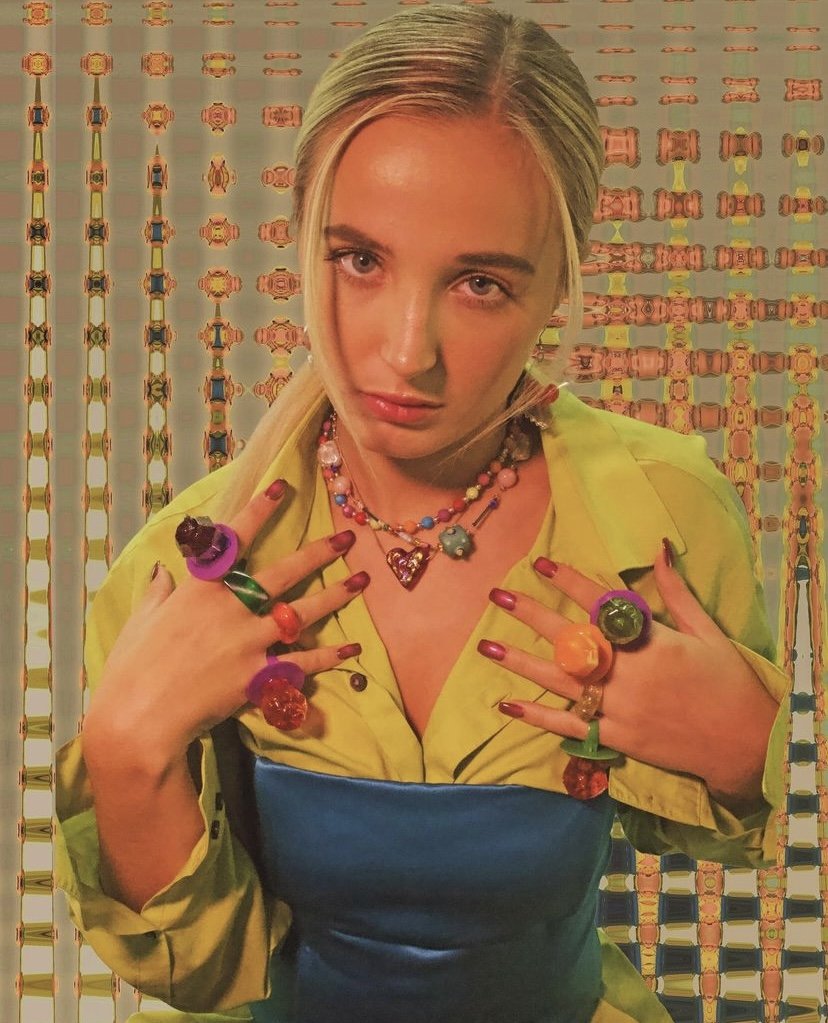 Woman with blonde hair wearing a yellow shirt and a colorful necklace, posing against a patterned background with various rings on her fingers.