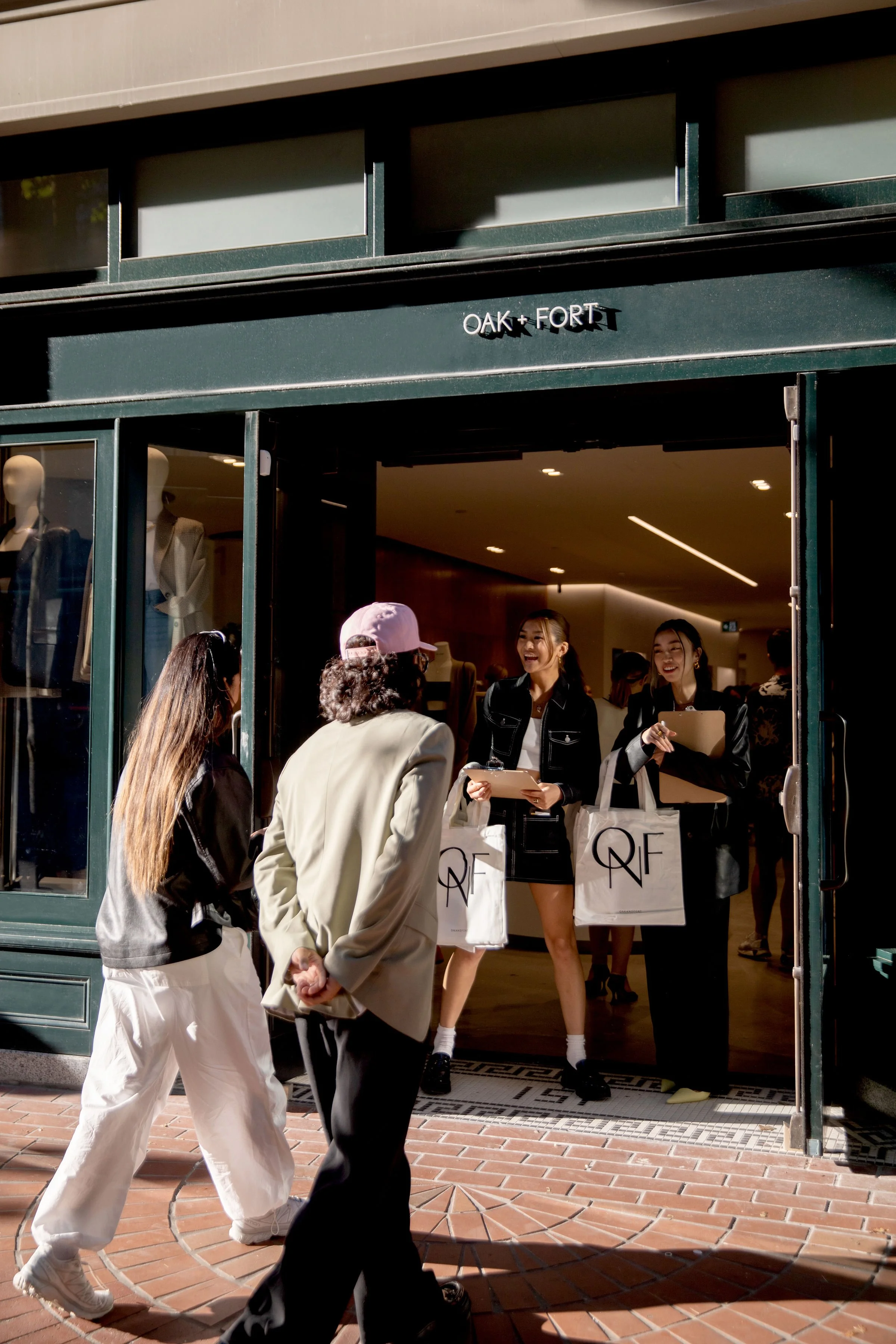 People shop at a retail store with a sign that says 'OAK + FORT'. Two employees inside the store are handing out shopping bags and talking to customers outside.