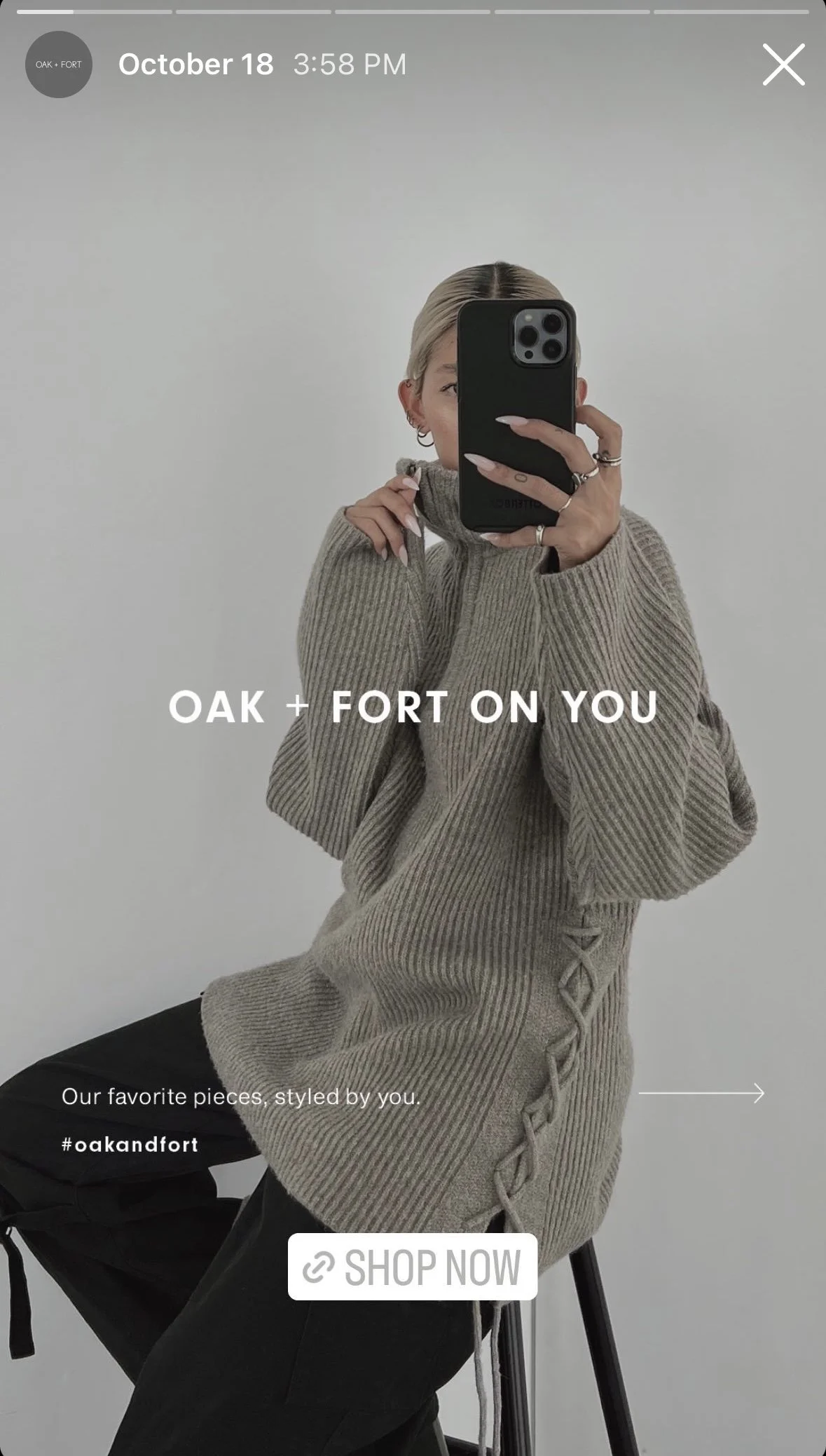 A woman wearing a beige ribbed knit sweater with a zipper detail on the side, sitting on a black stool in front of a plain white wall, taking a mirror selfie with her phone, with text overlay promoting Oak + Fort clothing.