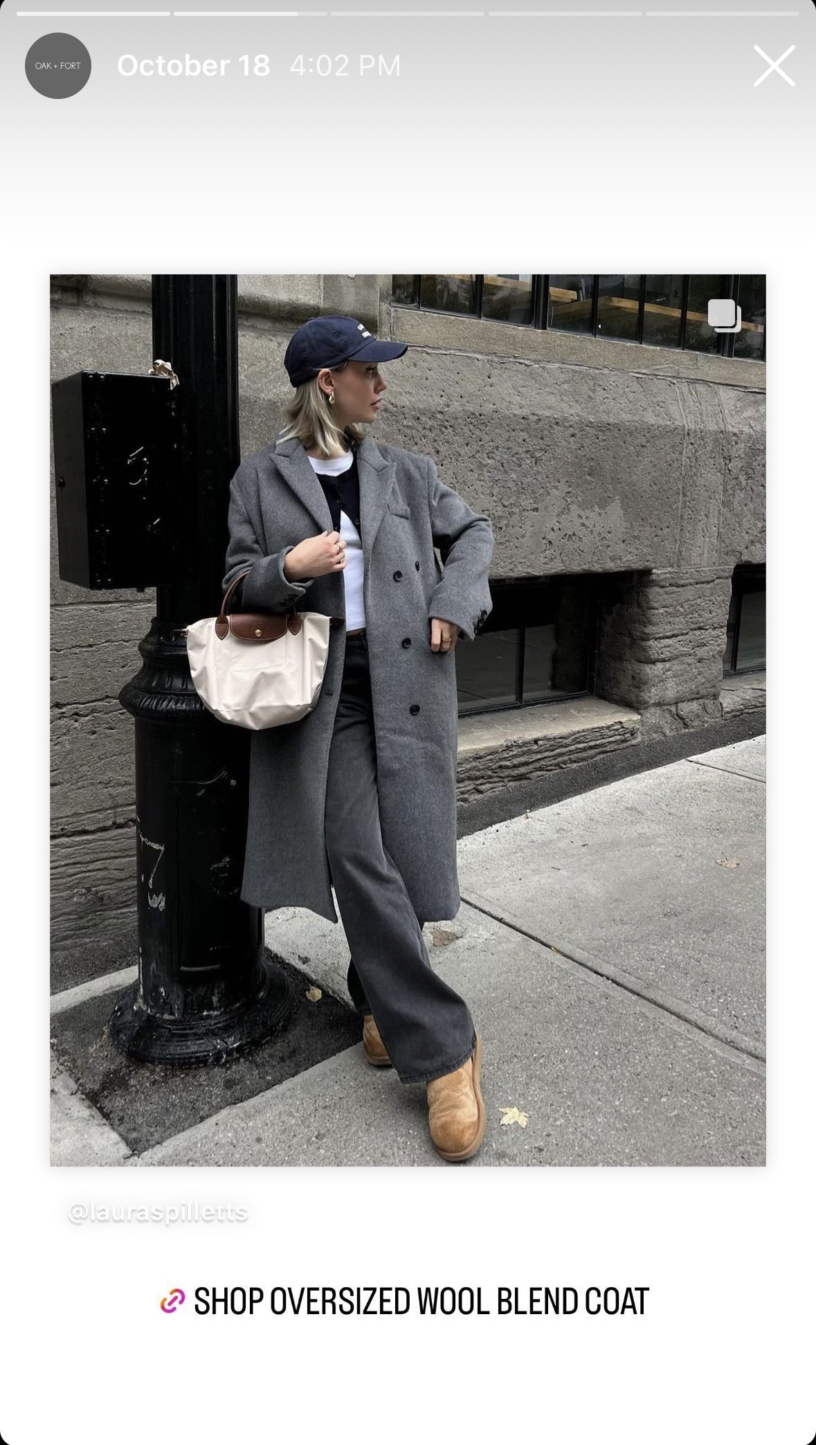 A woman is leaning against a black fire hydrant on a city sidewalk, dressed in a long gray wool coat, dark wide-leg jeans, a navy baseball cap, and tan boots, holding a beige tote bag. The background features a stone building with a window.
