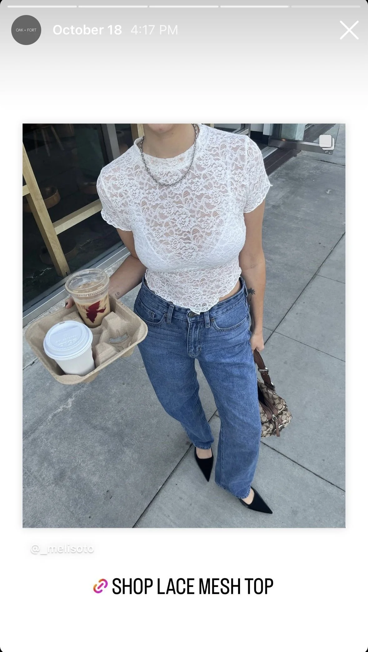 A woman wearing a white lace mesh top, blue jeans, and black pointed shoes. She is holding a tray with a coffee and a bottled drink, standing outside a storefront.