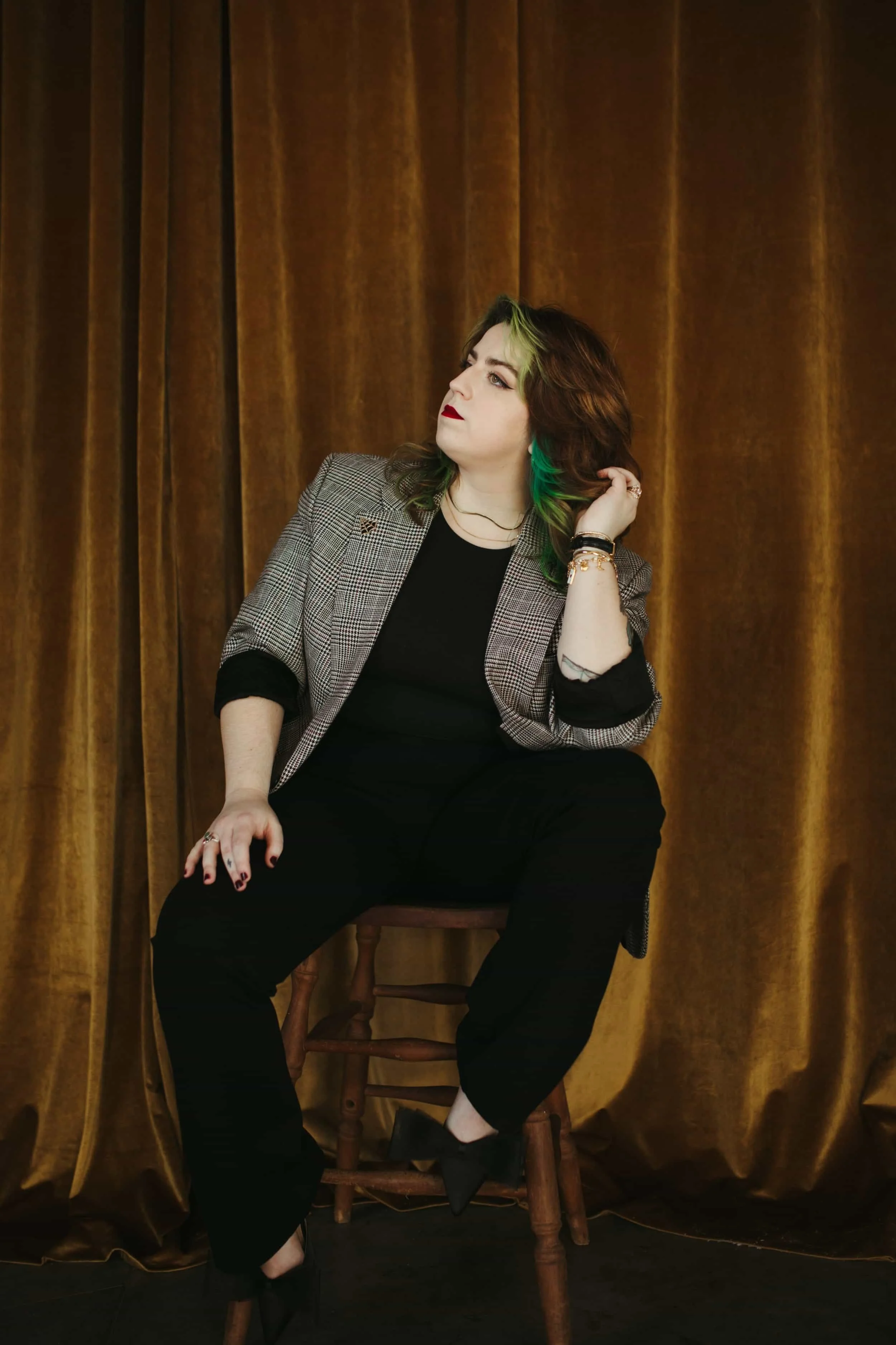 Kathleen sits on a wooden chair against a gold velvet curtain backdrop. She wears a checkered blazer, black top, and black pants, and gazes to her left with her hand in her hair.