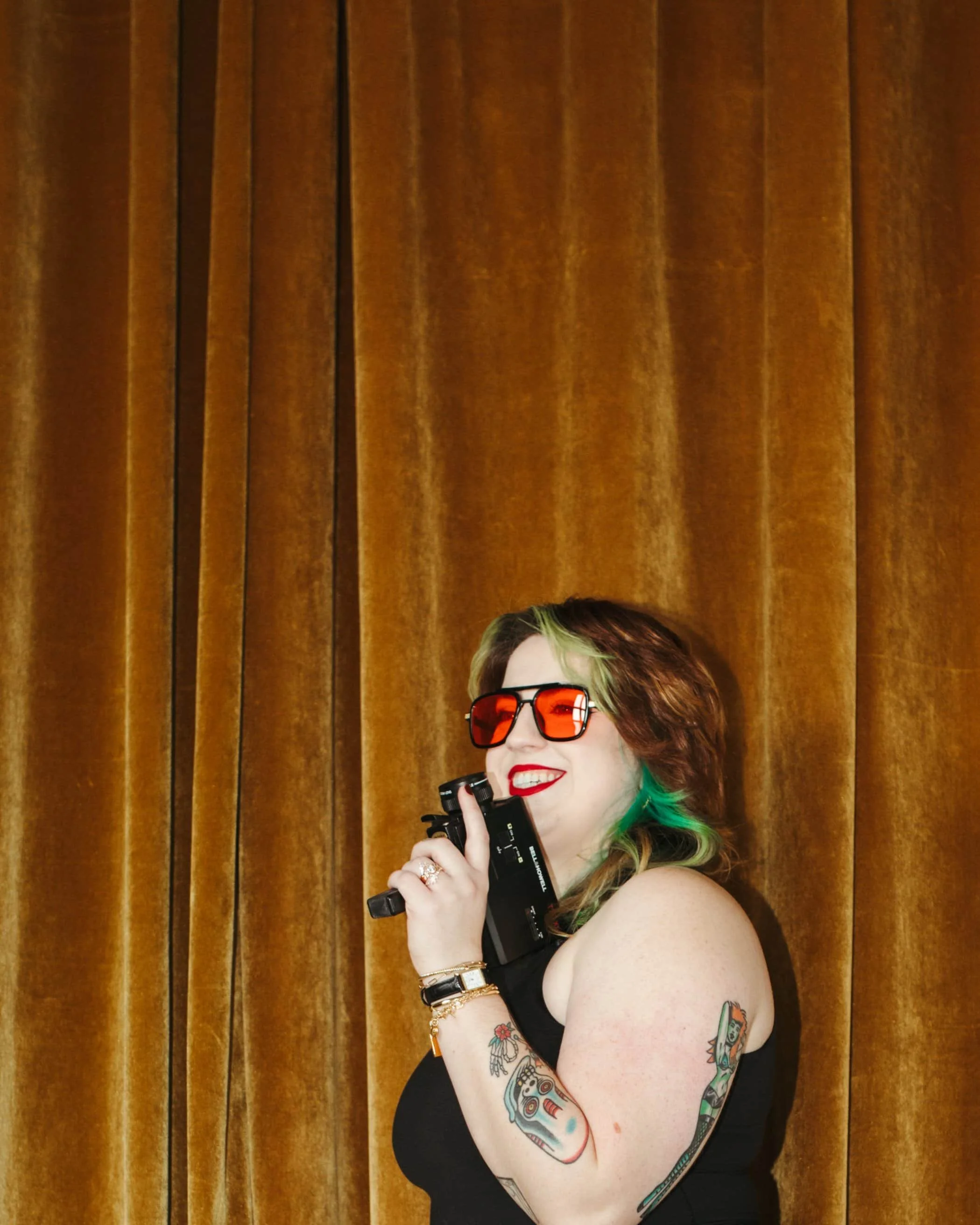 Kathleen stands in front of a golden velvet backdrop holding a super 8 film camera like a pistol. She is smiling away from the camera. She has green and blue hair and is wearing retro sunglasses with bright orange lenses.