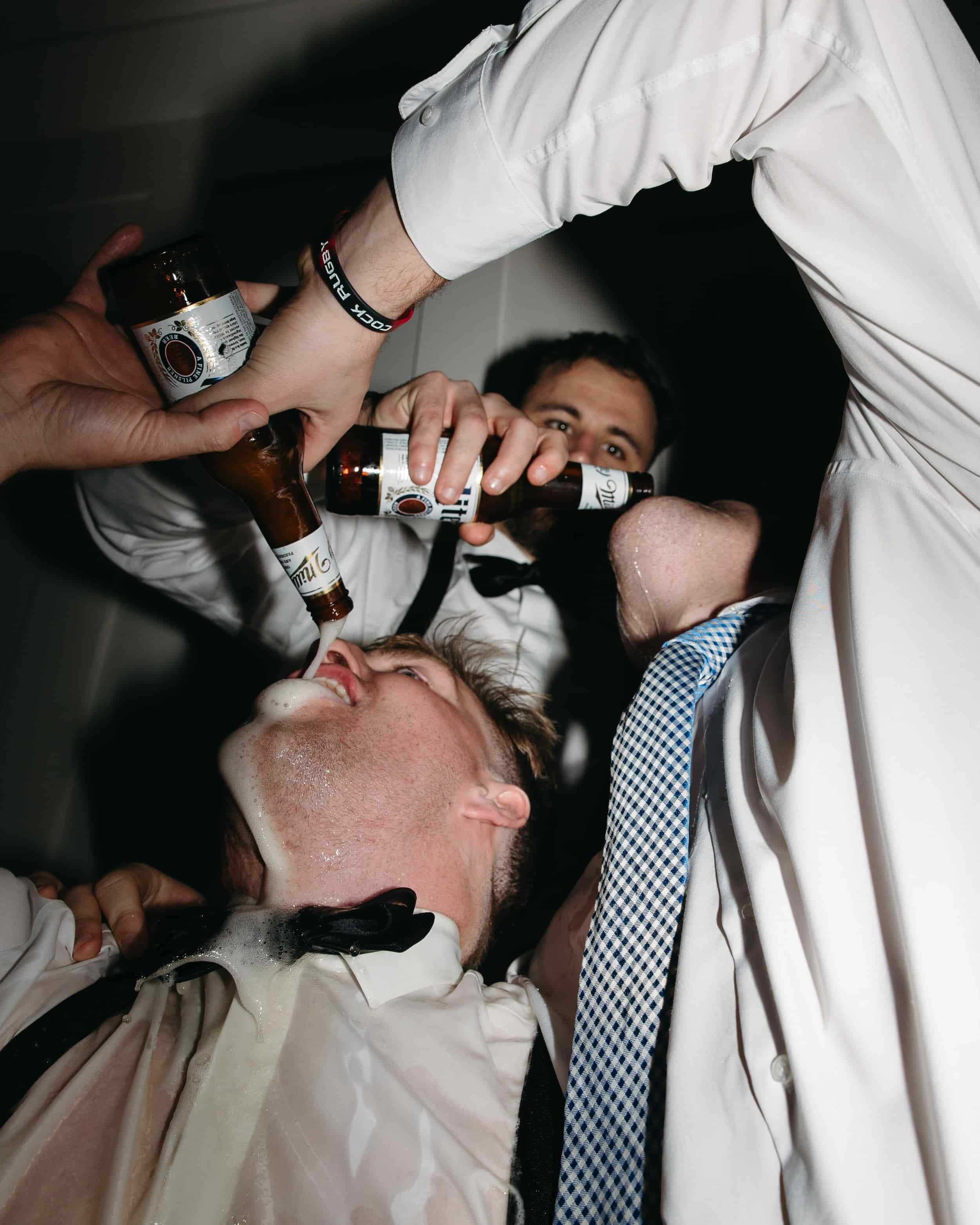 Groomsmen create a "beer waterfall" during a reception. Their shirts and faces are covered in beer. Photographed during a luxury Washington DC wedding at the National Museum of Women in the Arts. Kathleen is a luxury wedding photographer offering doc