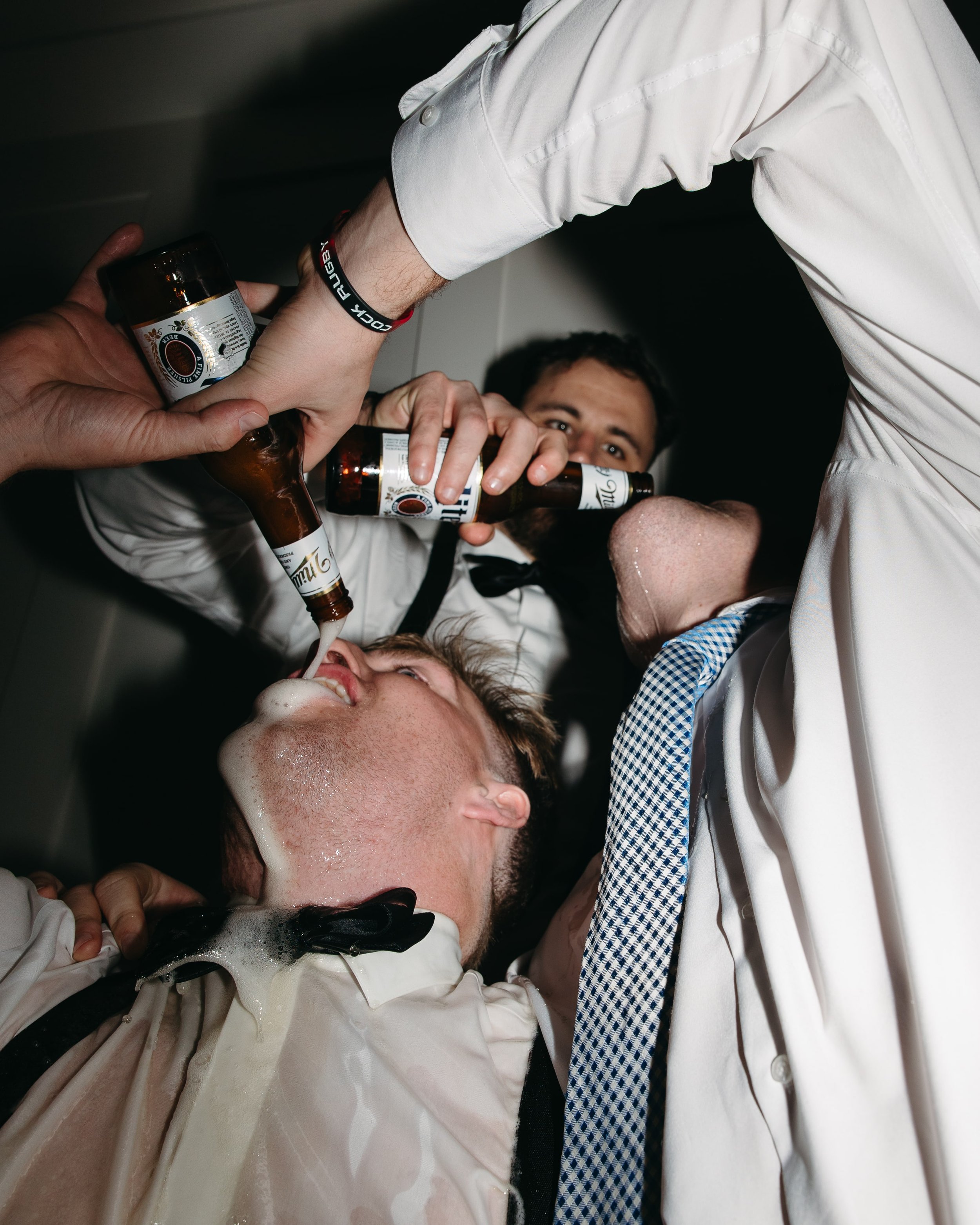 Editorial flash photography capturing a playful, raucous wedding moment by Kathleen Baker Photography. A groomsman lies back as friends pour a bottle of Miller High Life beer into his mouth, foam spilling down his shirt. This raw, unscripted scene hi