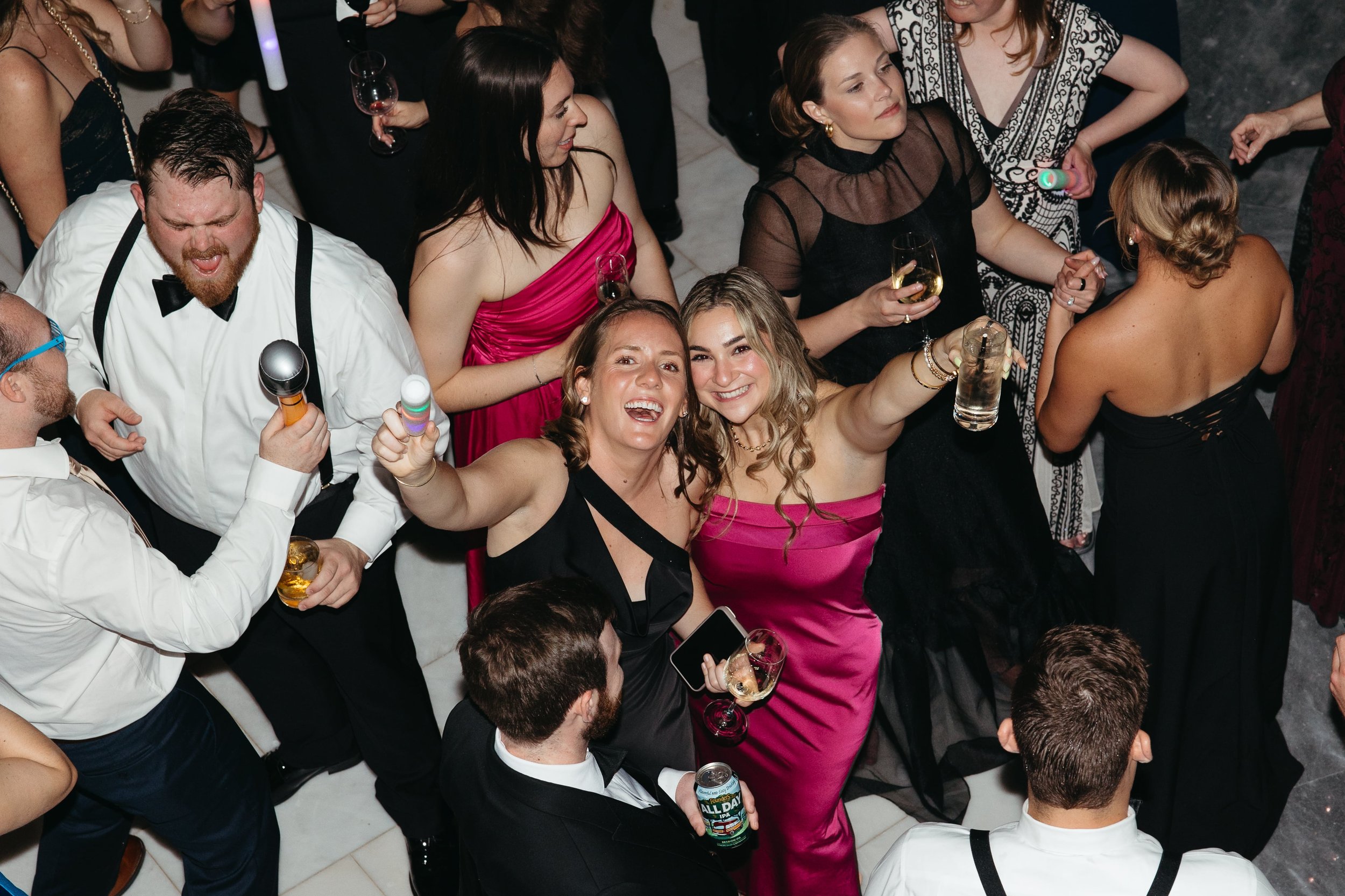 A joyful reception photograph by luxury wedding photographer Kathleen Baker, featuring guests in elegant black and jewel-toned gowns dancing and cheering. Two women in the center, one in a black dress and one in a bright pink satin gown, raise their