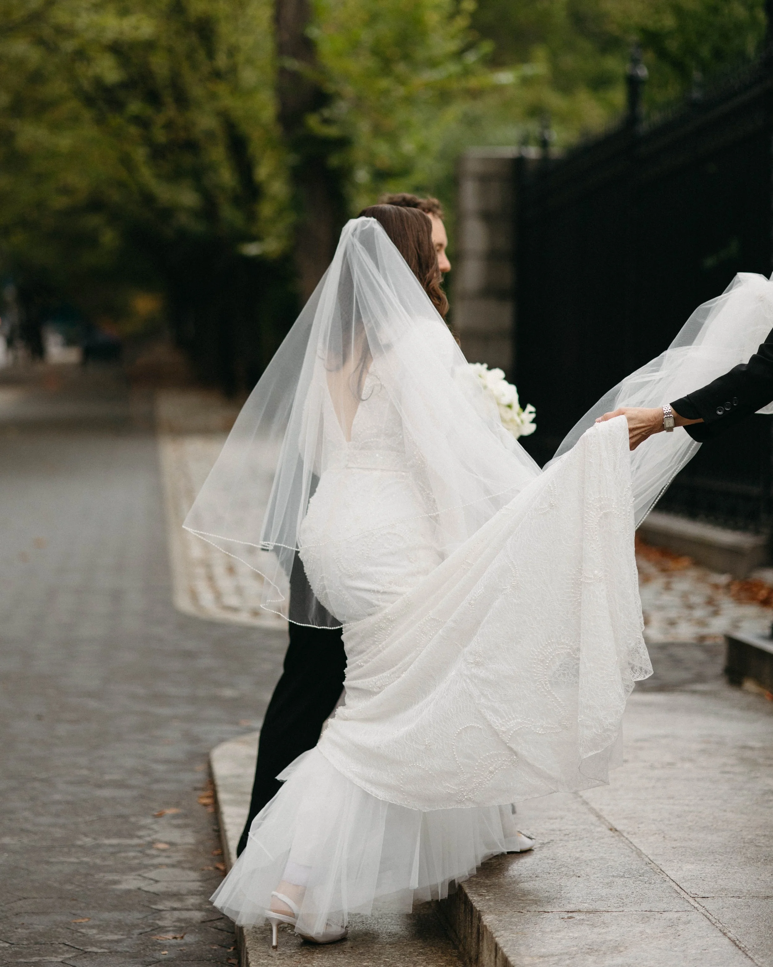 Bride walks ups two stone steps outside of a park in NYC. Her husband is holding her left hand, and the planner is holding her train out in front of her to keep it off of the slightly damp ground. She has a cathedral veil and beaded wedding gown.