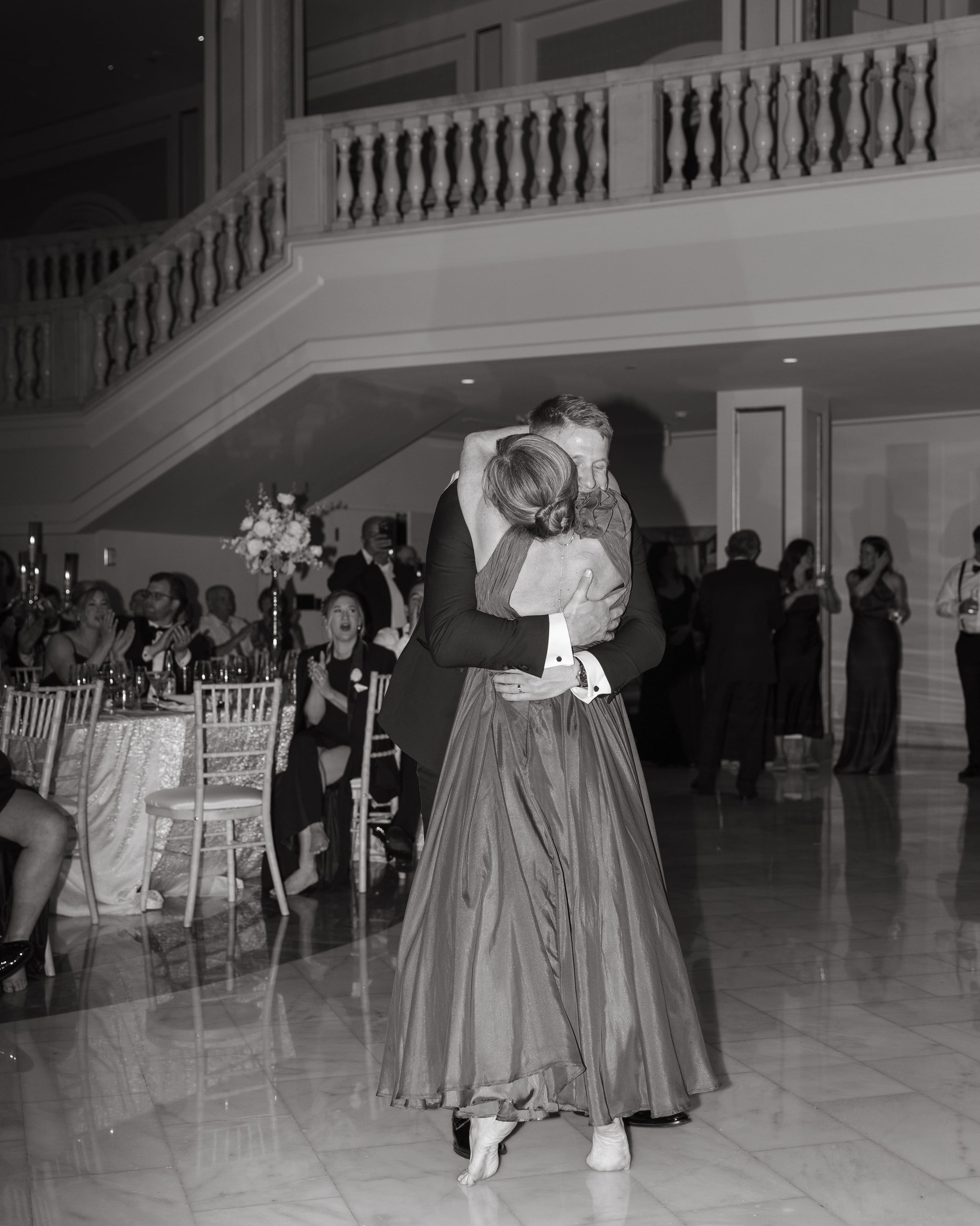 Documentary black and white photo by Kathleen Baker Photography of an emotional mother-son dance at an elegant Washington DC wedding reception. The groom, dressed in a sharp tuxedo, lifts his mother in a flowing satin gown off the marble dance floor