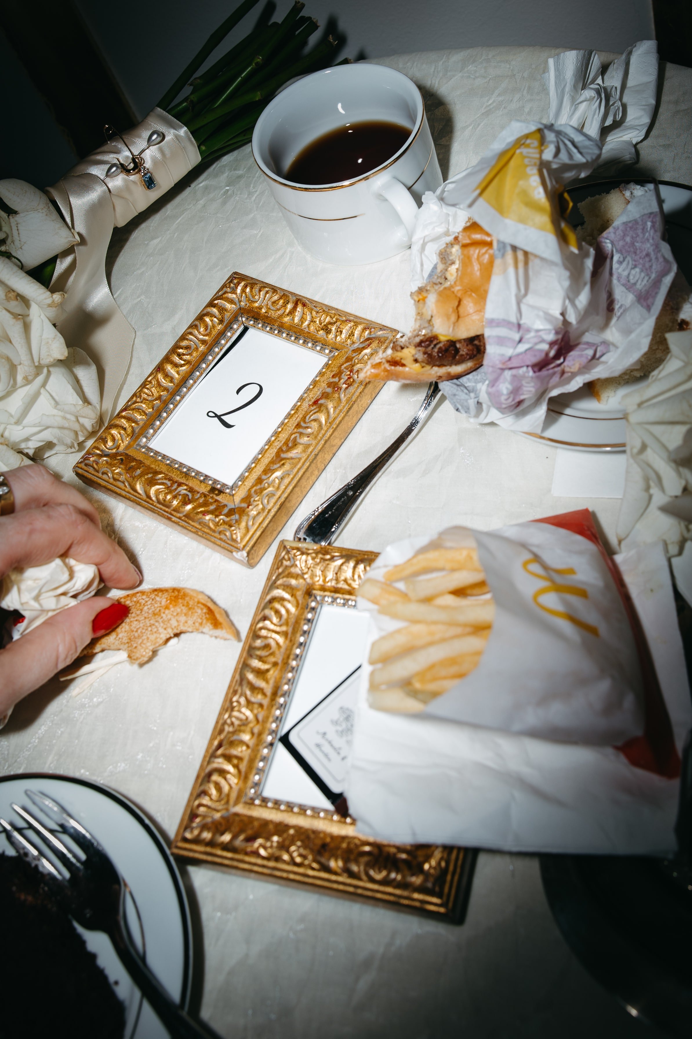 A detail-rich editorial photo by DC and NYC wedding photographer Kathleen Baker of a late-night snack vignette at a wedding reception. On a white linen-covered table sit a bouquet of white roses, a fine china coffee cup, and McDonald’s cheeseburger a